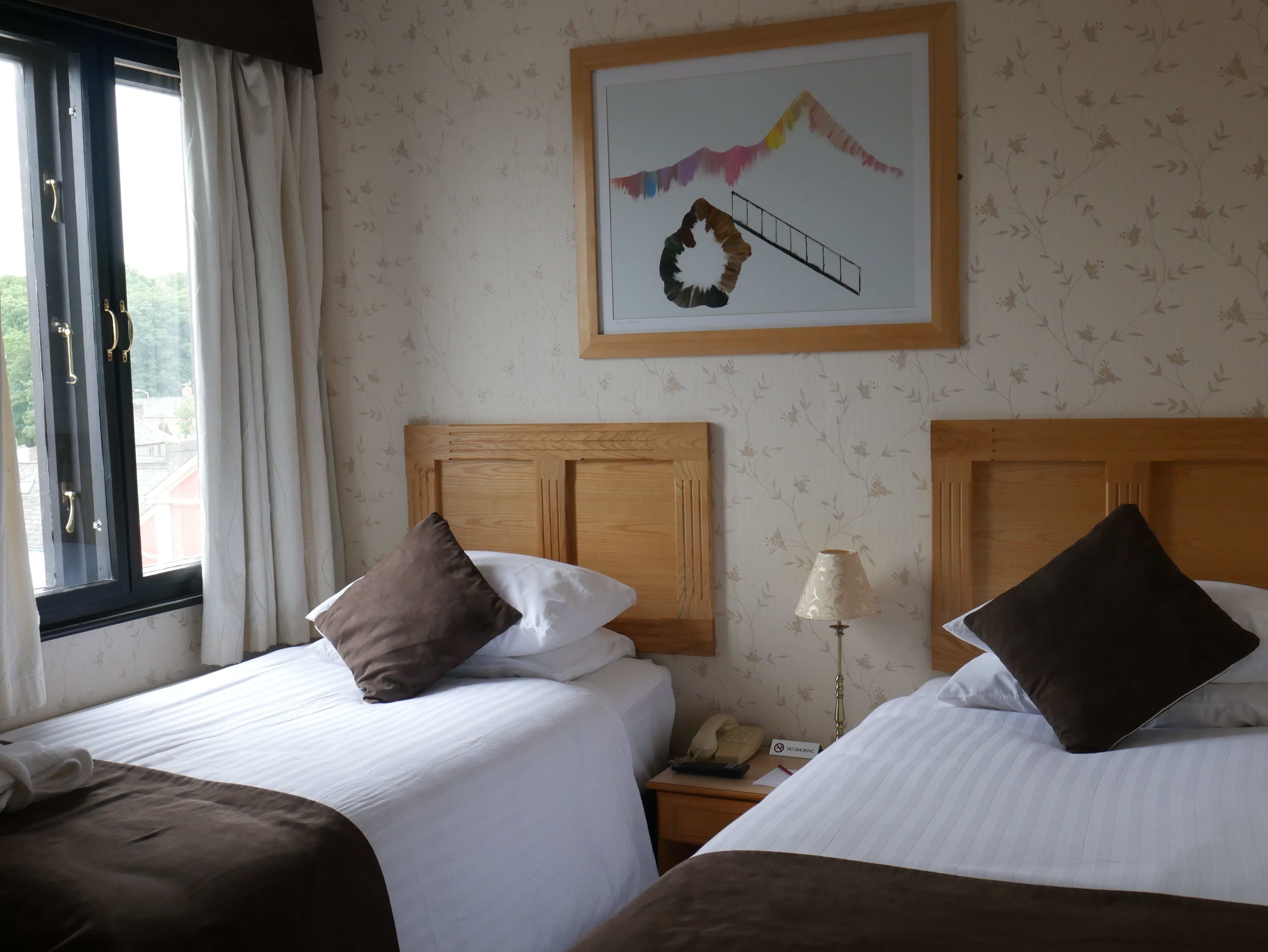 Habitación de hotel con dos camas, una mesita de noche con un teléfono y un cartel de 'No Fumar', una ventana con cortinas y un papel tapiz floral.