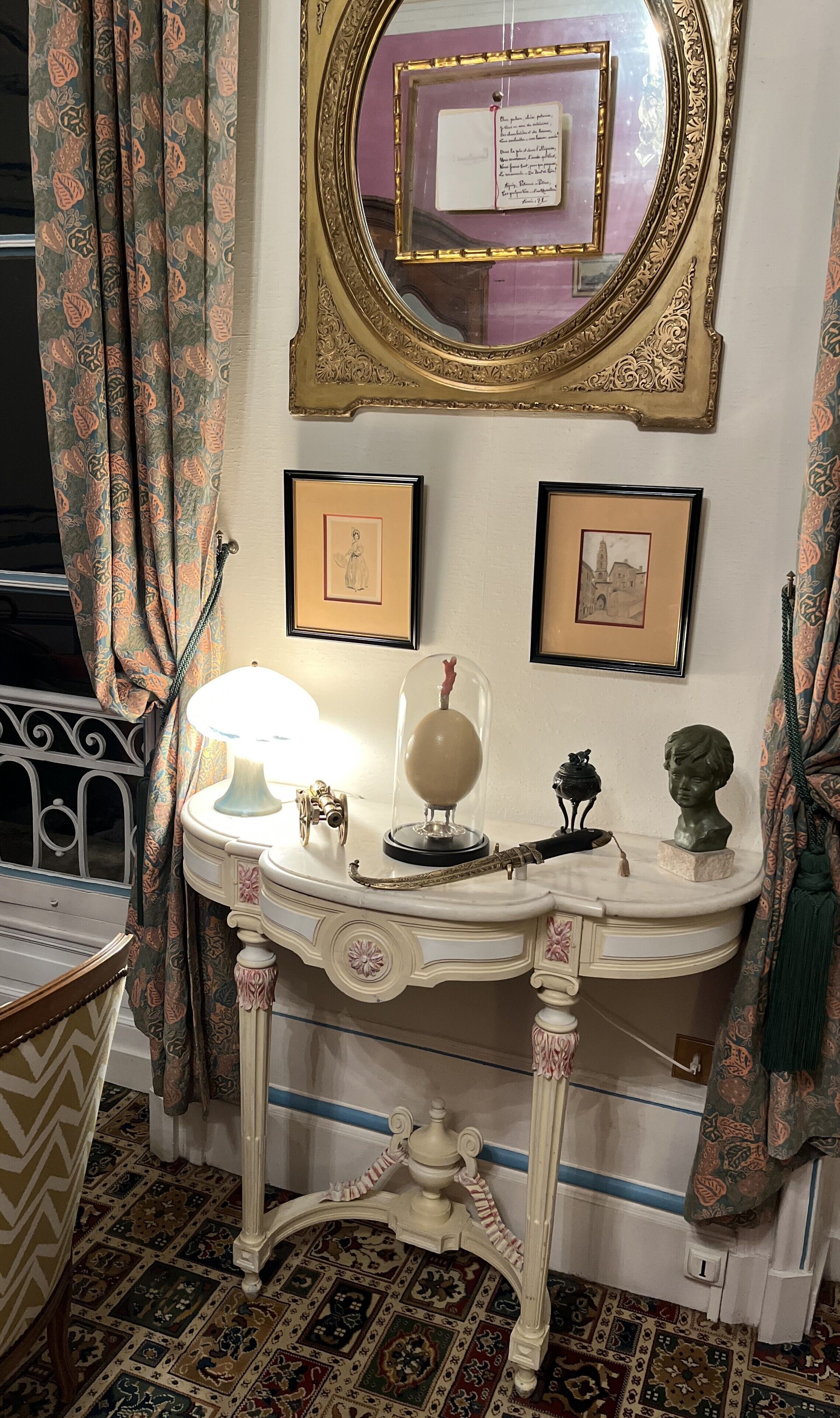 A room with a decorative table, a mirror, framed pictures, and a lamp.