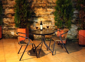 Un espace extérieur aménagé avec un mur en pierre, deux fauteuils en osier, une table ronde et une bouteille de vin.