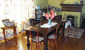A dining room with a wooden table, chairs, a vase with flowers, a fireplace, and a bookshelf. - La Casona del Rio (Trelew)