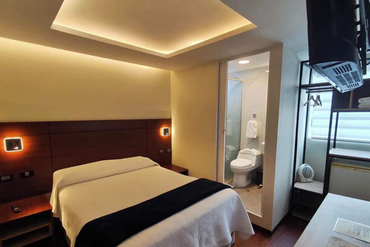 Ein Hotelzimmer mit Bett, Nachttisch und einem kleinen Schreibtisch mit Stuhl. Ein Fenster bietet einen Ausblick ins Freie.