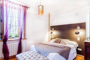 Une chambre à coucher comprenant un lit, une table de chevet, un luminaire suspendu et une fenêtre avec des rideaux.