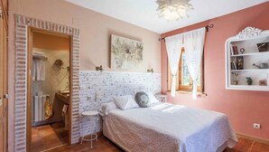 A bedroom with a white brick headboard, a bed with white bedding, a white shelf with books and decorative items, and a window with white curtains. - El Torreón de Navacerrada (Navacerrada)