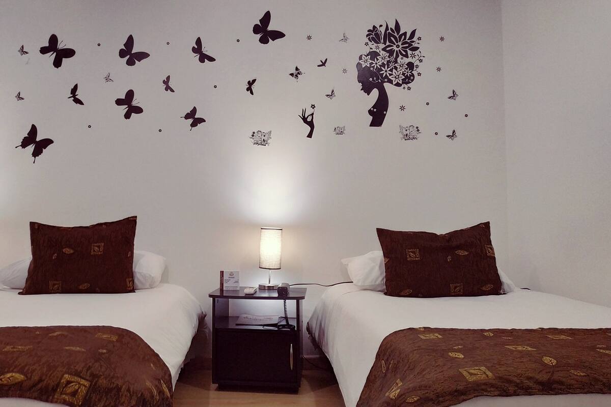 Quarto de hotel com duas camas, cada uma com roupa de cama e almofadas castanhas, uma mesa de cabeceira com abajur e decoração na parede com borboletas e uma árvore.