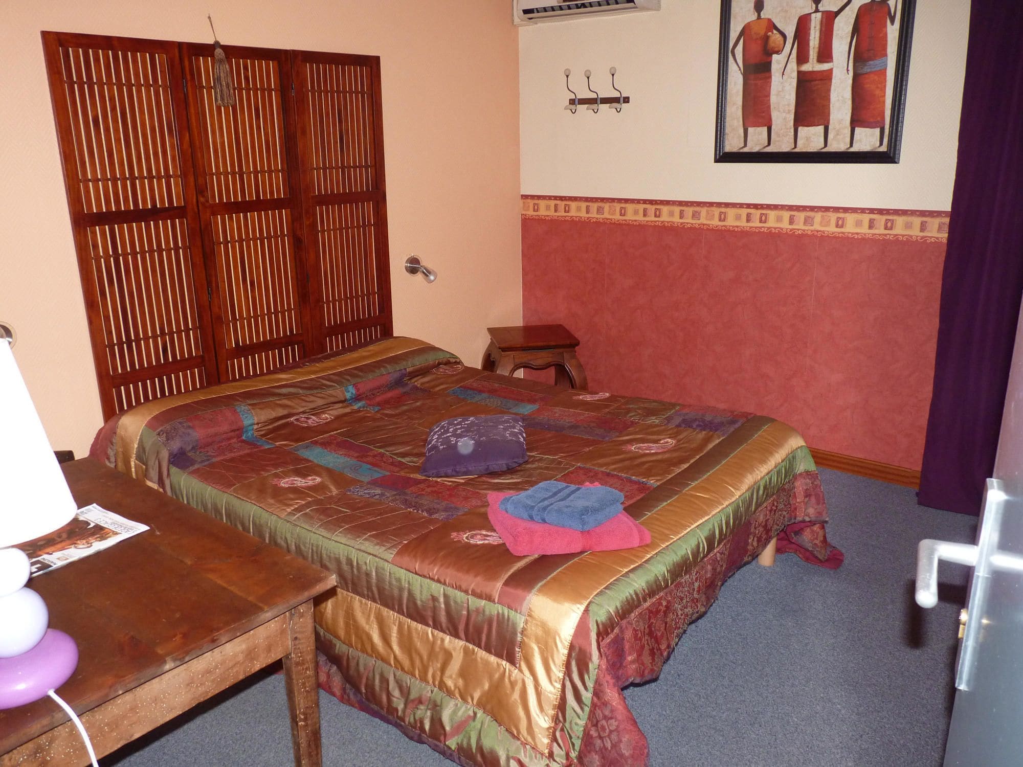A bedroom with a bed, a wooden desk, a small stool, a wall-mounted air conditioning unit, and a framed picture.