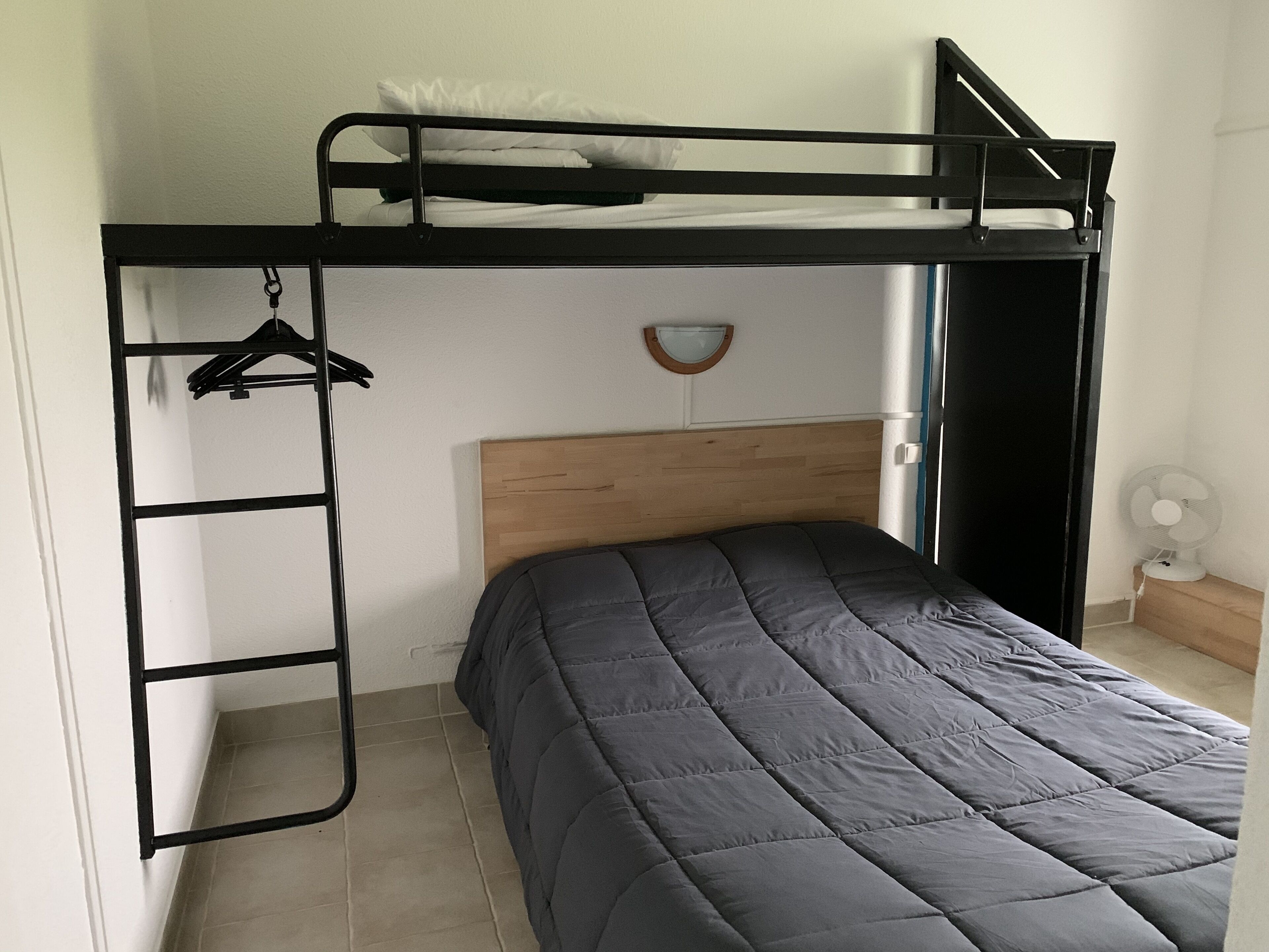 A bunk bed with a wooden headboard, a single bed below, a wall-mounted shelf, and a small fan on the floor.