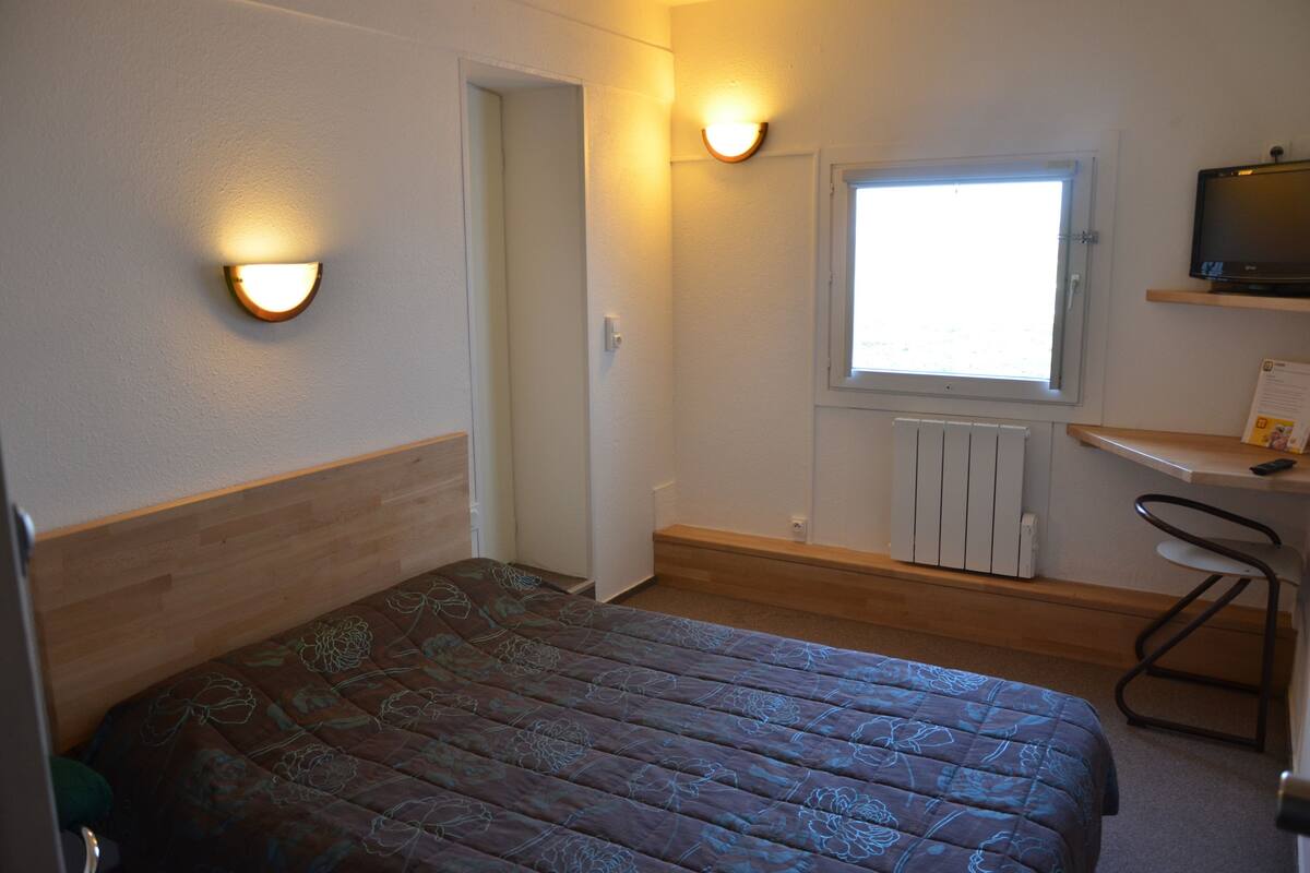 A small, well-lit room with a bed, a desk, and a window.