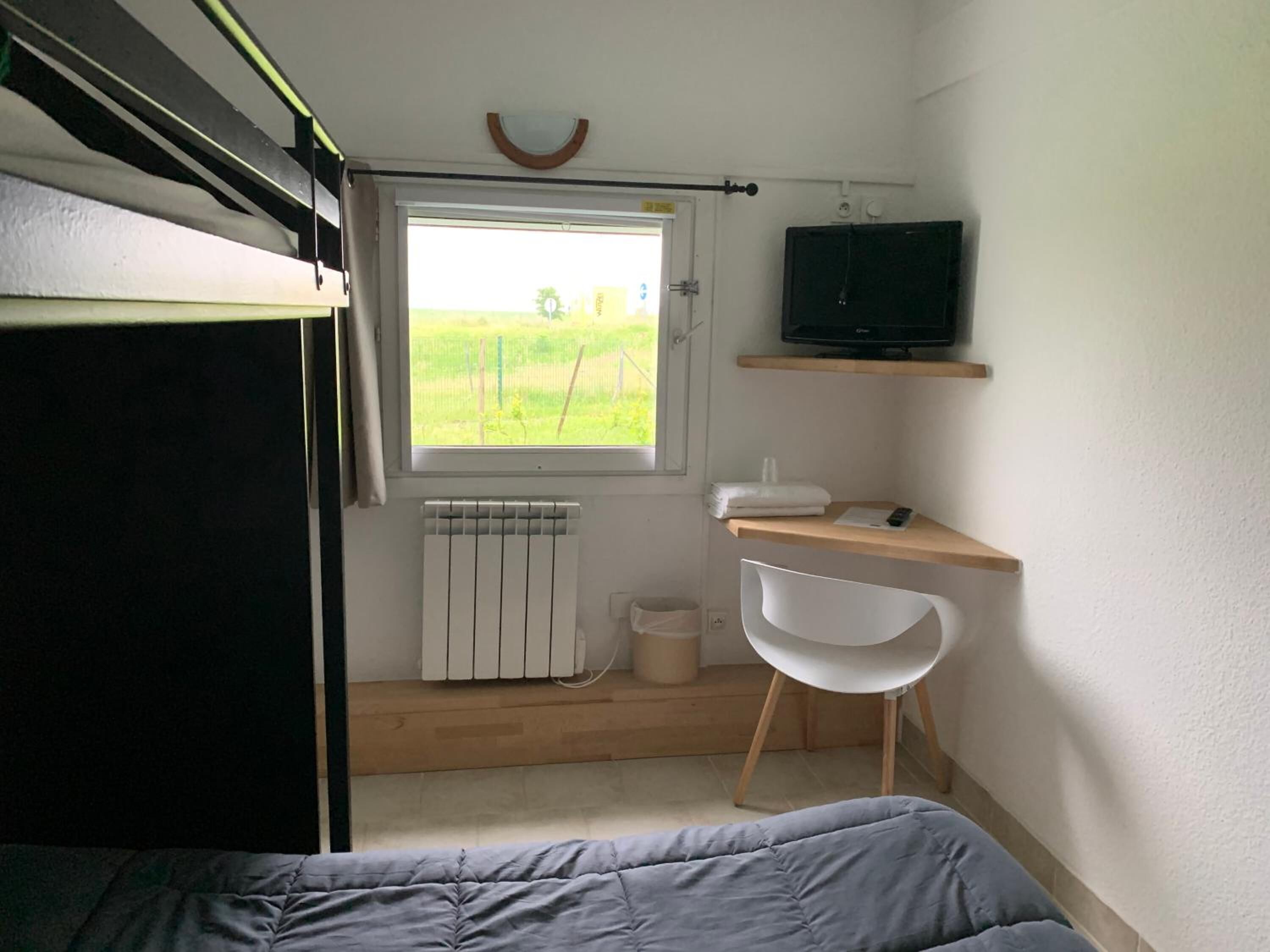Une chambre à coucher de taille modeste, comprenant un lit, une fenêtre donnant sur un espace verdoyant, un radiateur, un bureau avec une chaise et un téléviseur posé sur une étagère.