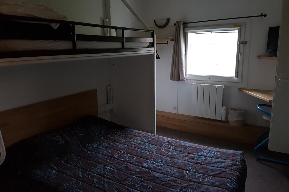 Una habitación con literas, una cama individual con colcha estampada, una ventana con cortinas, un radiador y un estante con un televisor.