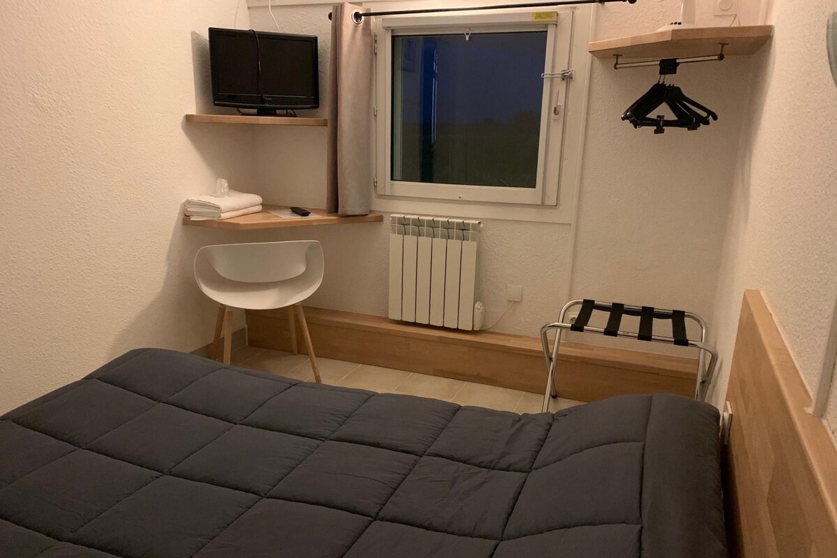 Una habitación pequeña con cama, una silla, un escritorio, ventana y radiador.