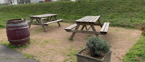 Un área de picnic con dos mesas de madera y un barril, rodeada de césped y una jardinera de concreto con un arbusto.