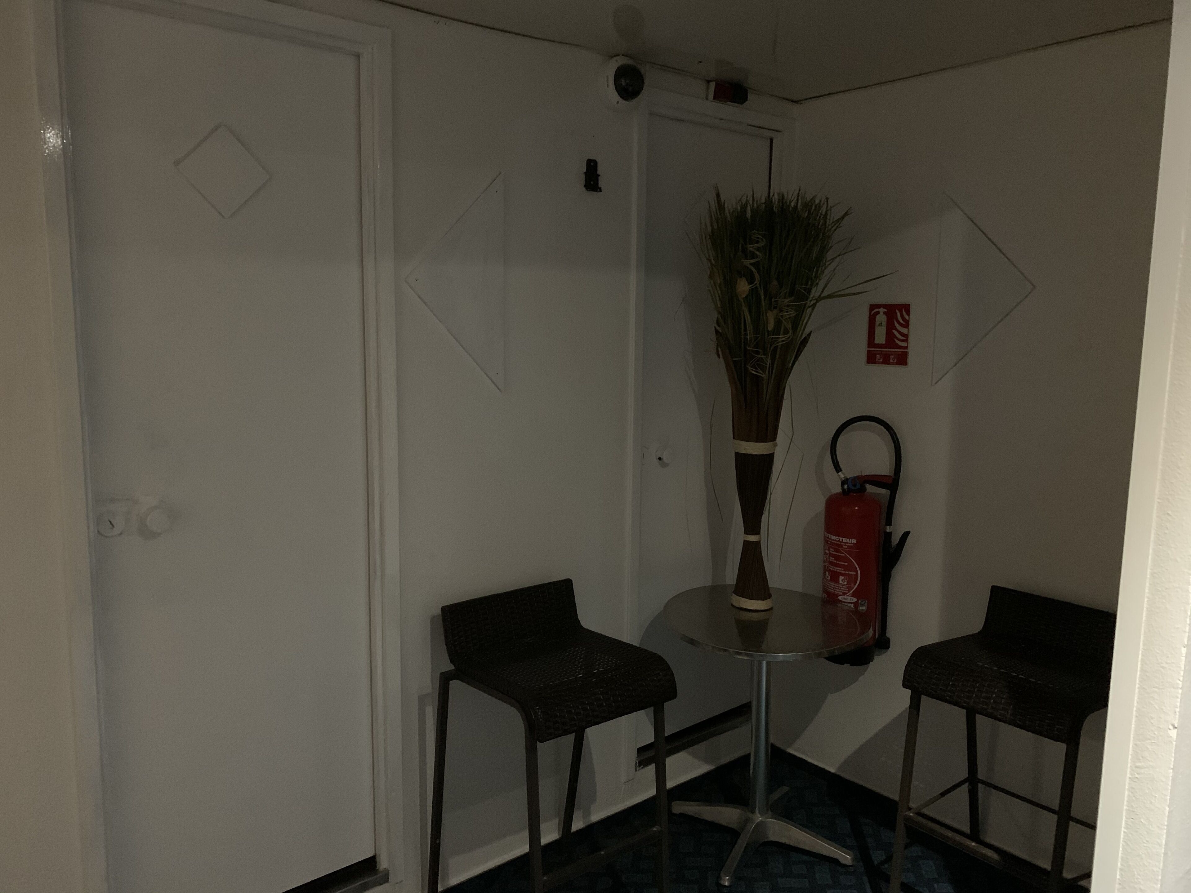 Um quarto pequeno com uma porta branca, uma mesinha com um vaso de plantas, duas cadeiras e um extintor de incêndio na parede.