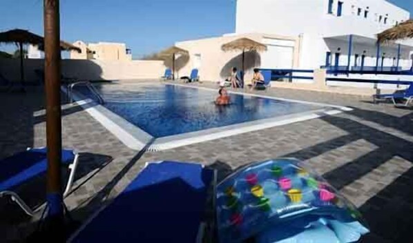 A swimming pool with lounge chairs and umbrellas, surrounded by a white building with blue accents. - Rooms Mary (Santorini)