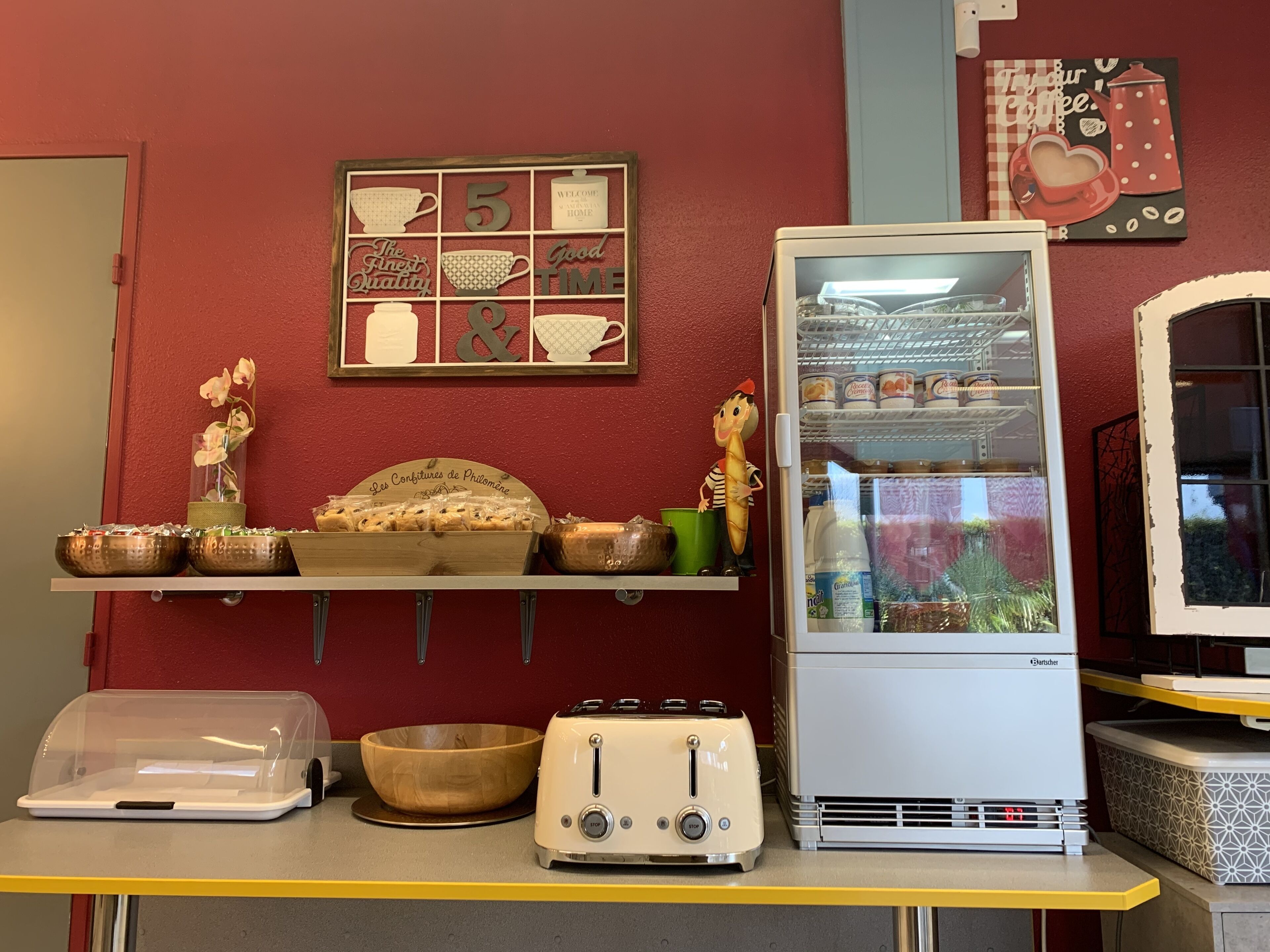 A kitchen area with a refrigerator, a toaster, a wooden bowl, and a shelf with baked goods.