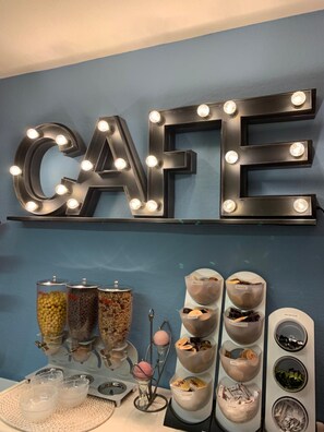 A cafe with illuminated letters spelling "CAFE" on the wall, a beverage dispenser, and a display of various drinks and snacks. - Europe Hôtel (Varennes-Vauzelles)