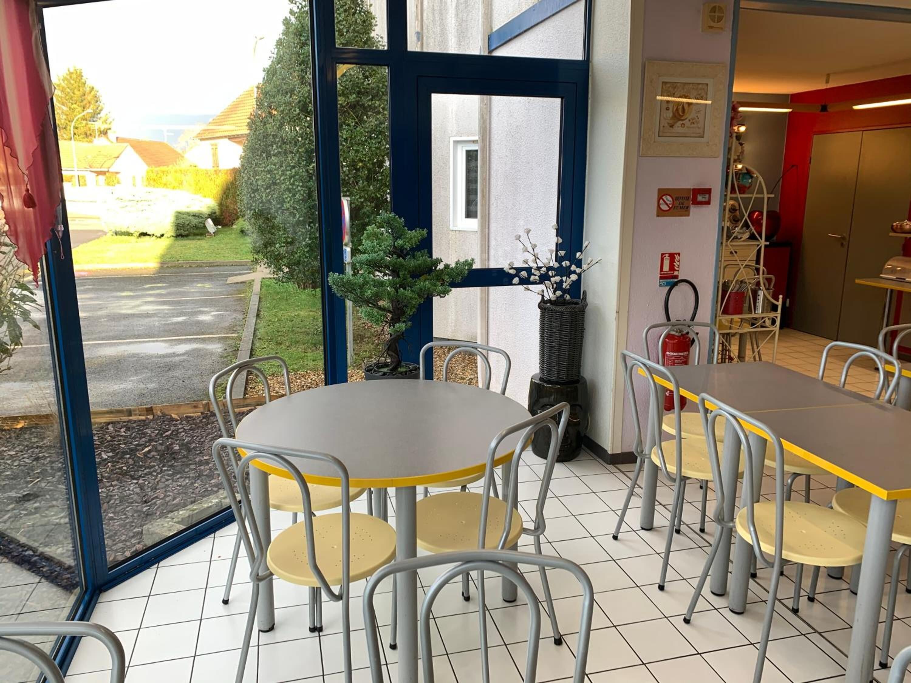 Een eetruimte binnenshuis met tafels en stoelen, met uitzicht op de straat en een brandblusser aan de muur.