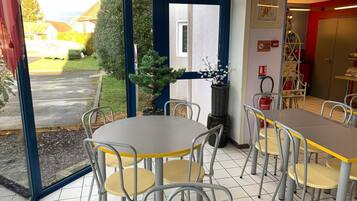 Een eetruimte binnenshuis met tafels en stoelen, met uitzicht op de straat en een brandblusser aan de muur.