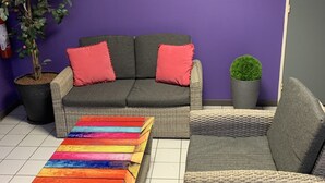 Een kamer met een paarse wand, een stadslandschap schilderij, een rieten bank met rode kussens, een kleurrijke gestreepte poef en een potplant.
