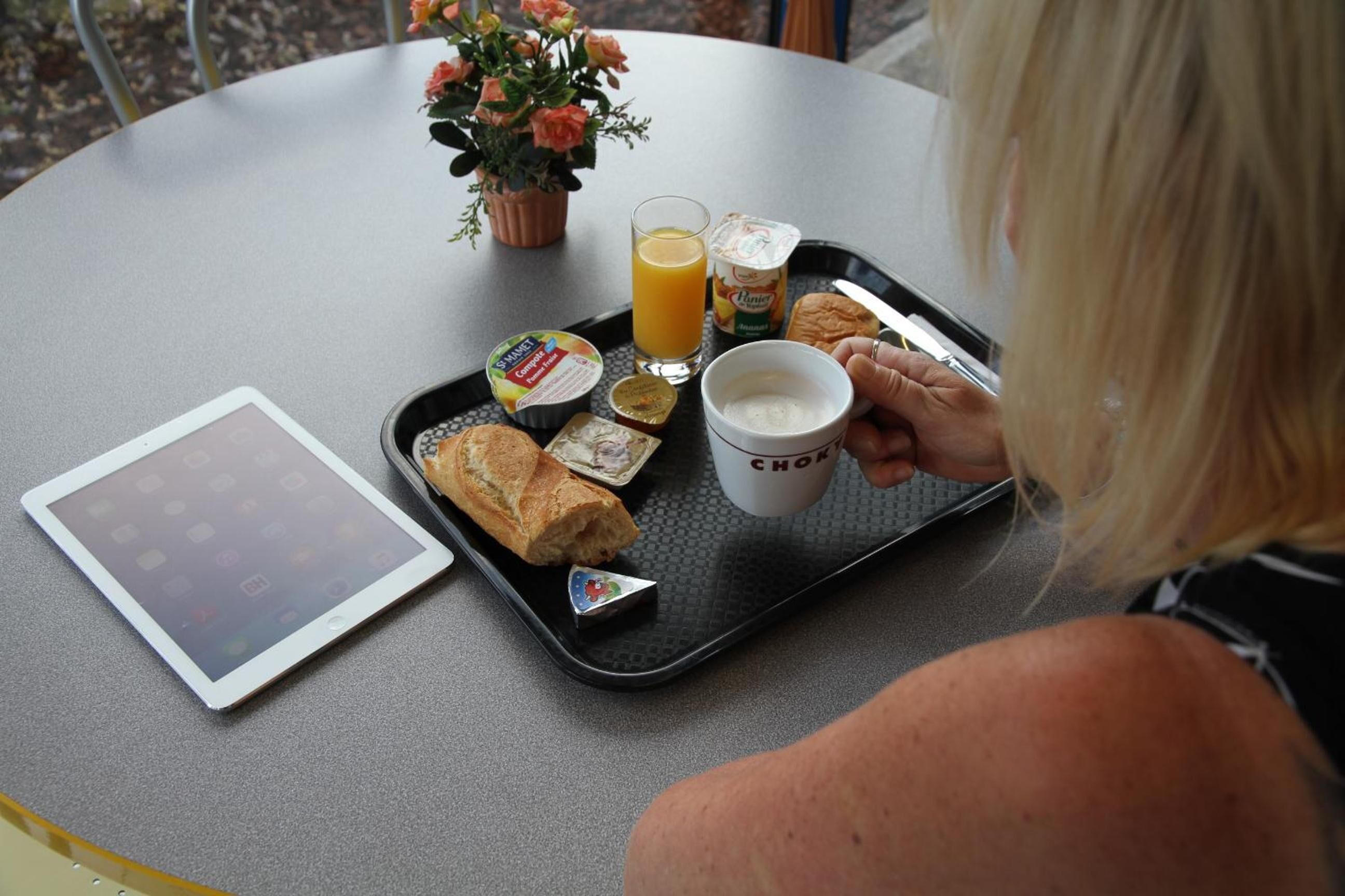 Ein Tablett mit Frühstücksartikeln, darunter Toast, Saft und eine Kaffeetasse, steht auf einem Tisch mit einem Tablet und einem Blumentopf.