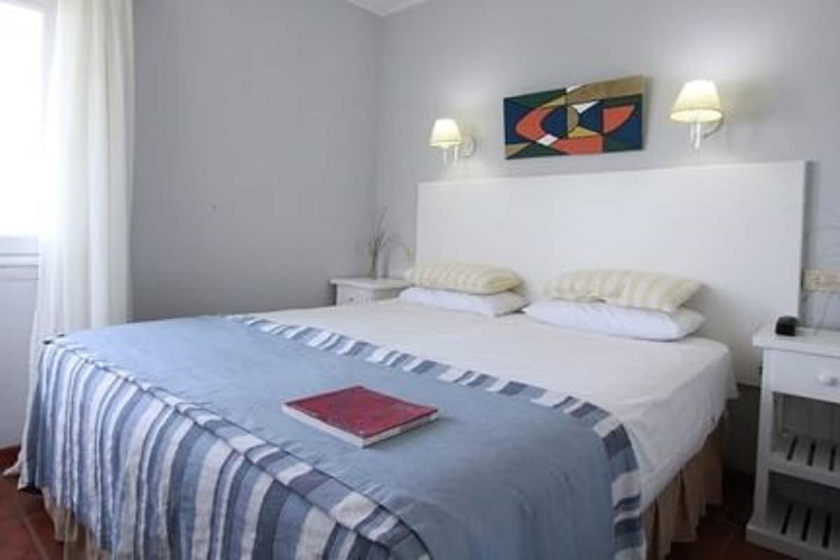 A bedroom with a bed, two bedside tables, a nightstand, a window with curtains, and a wall-mounted abstract painting.