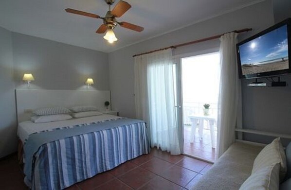 A bedroom with a bed, a ceiling fan, a TV, and a balcony with a view.