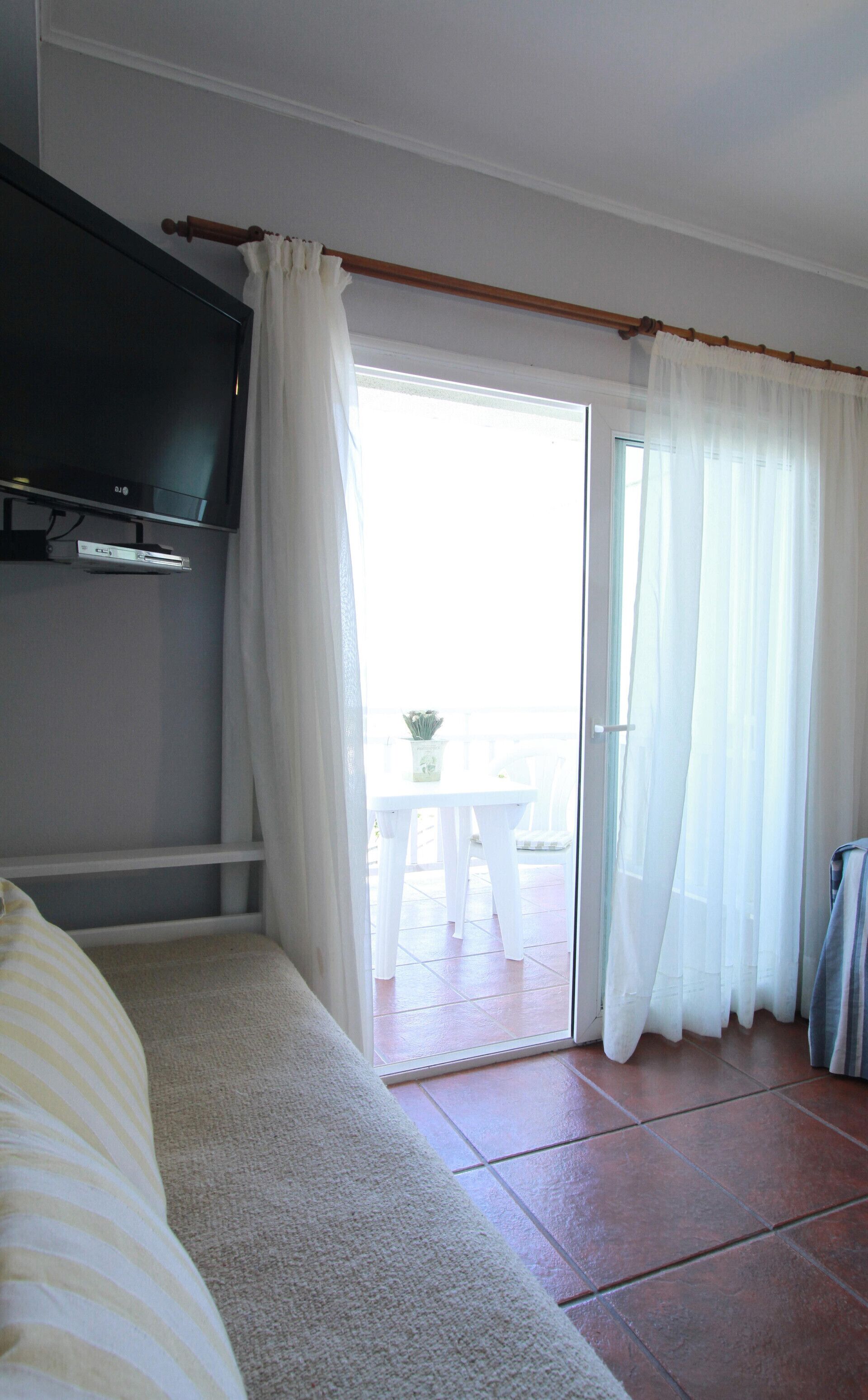Quarto com televisão, sofá, pequena mesa com planta e porta de vidro deslizante para área externa.