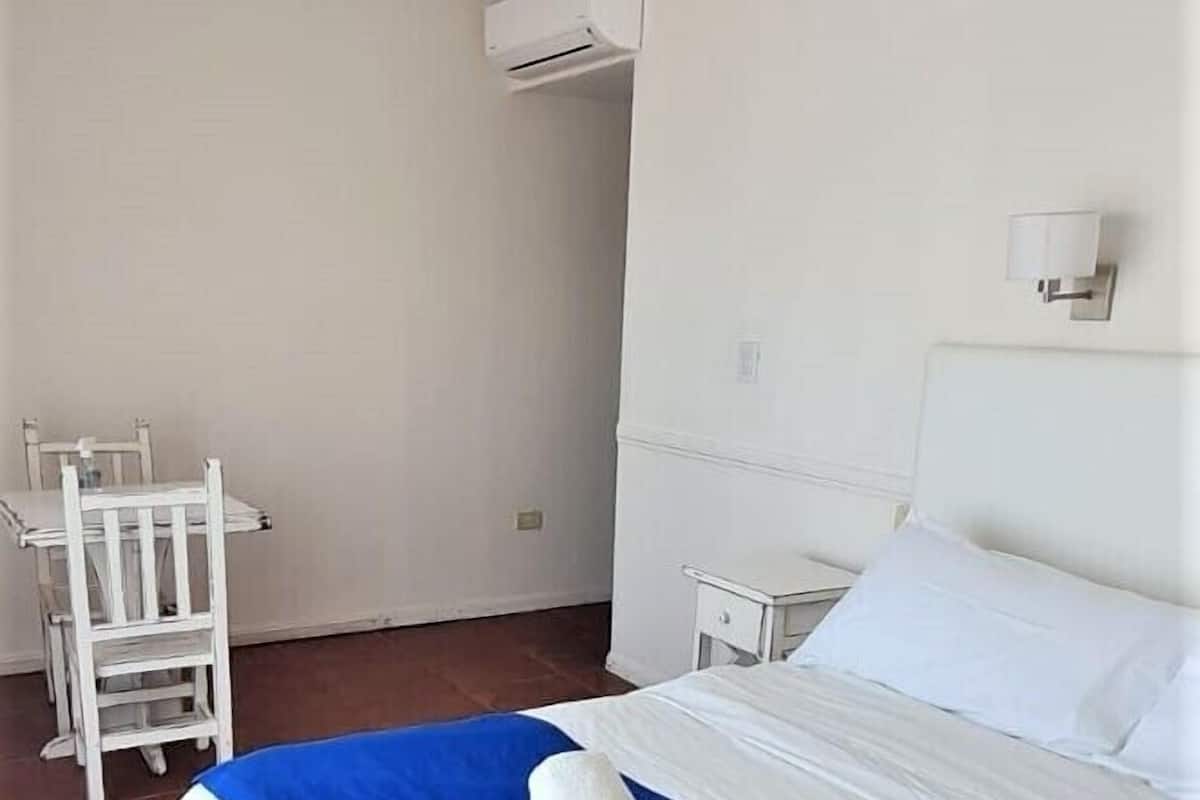 A hotel room with a bed, white walls, a ceiling fan, an air conditioning unit, a nightstand, a bedside table, and a white chair.
