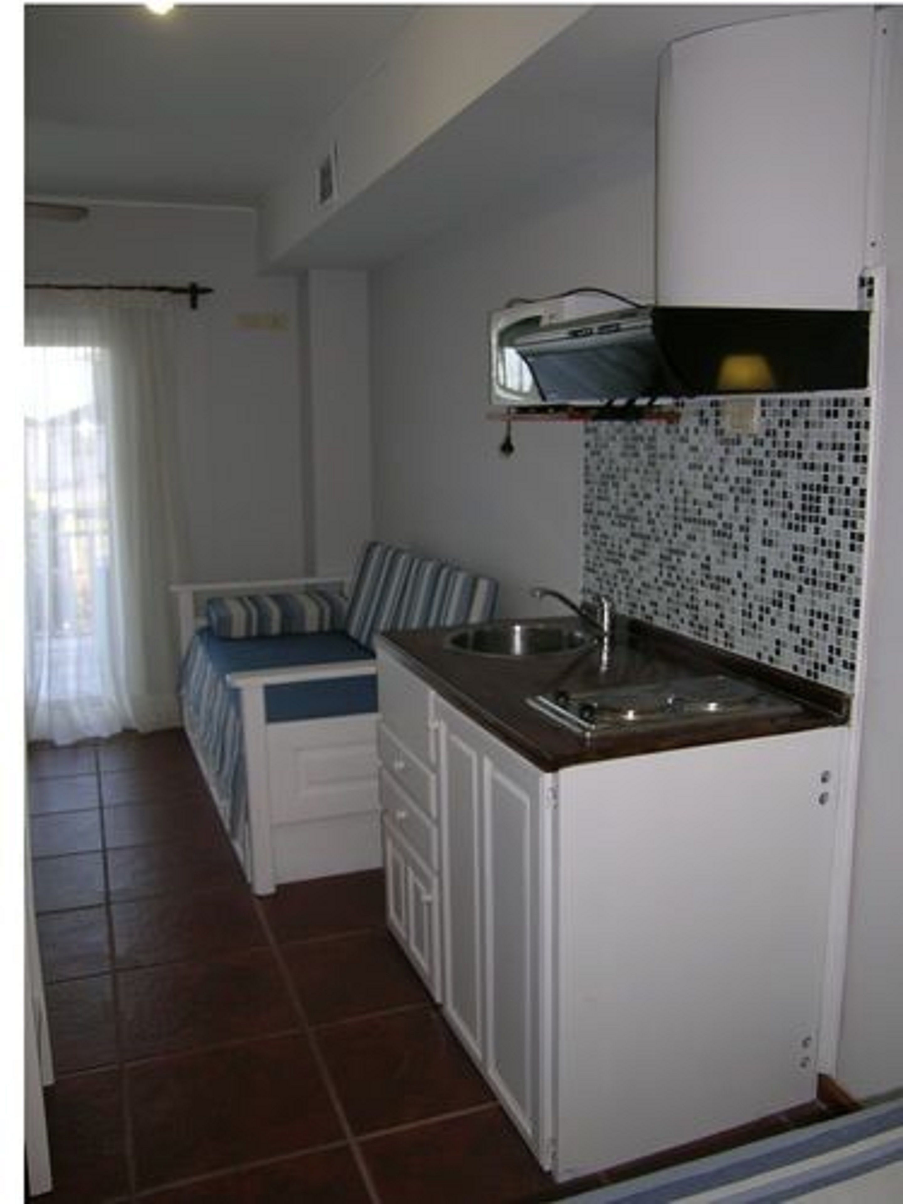 Cocina compacta con lavamanos, microondas y una pequeña encimera. Hay una cama con almohada a rayas y una ventana con cortinas.