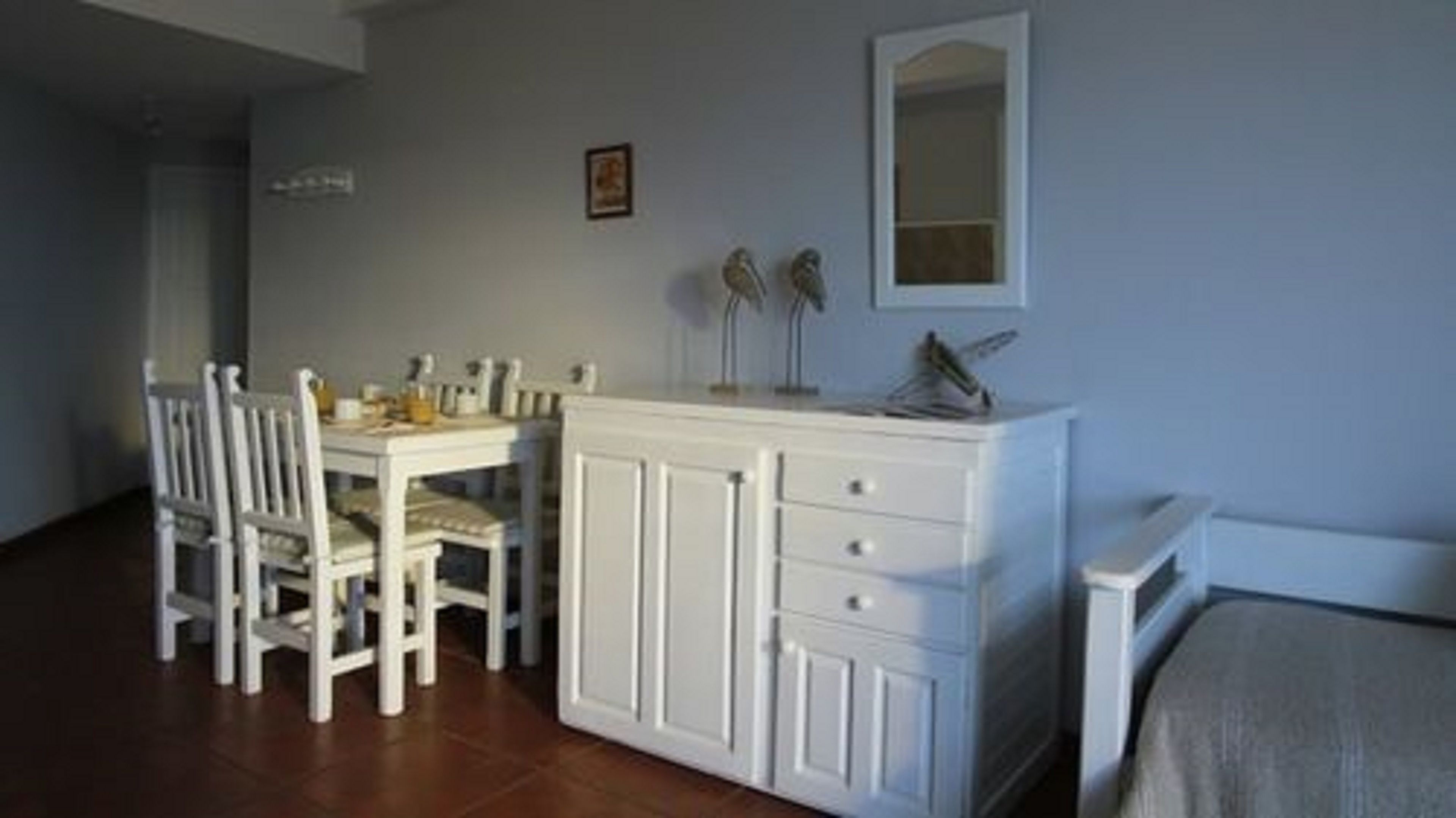 Un coin repas avec une table et des chaises blanches, un meuble blanc avec des tiroirs et un miroir au mur.