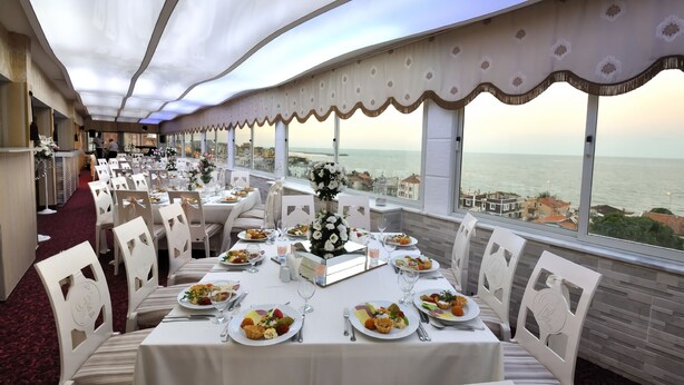 A dining area with tables set for a meal, large windows offering a view of the sea, and a high ceiling with a patterned design. - Grand Atakum Hotel (Atakum)