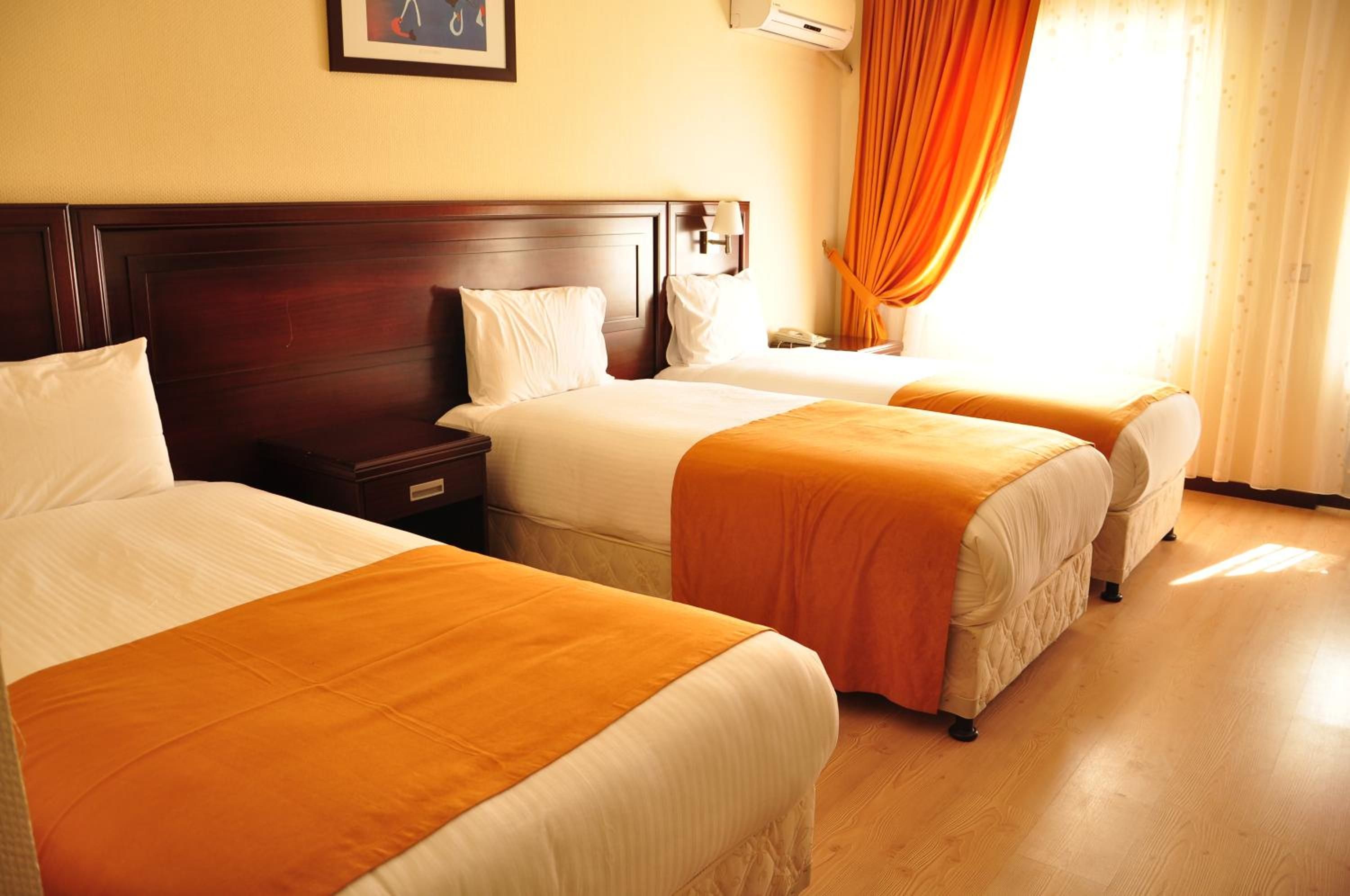 Une chambre d’hôtel avec trois lits simples, une tête de lit en bois, une table de chevet avec une lampe et une fenêtre avec des rideaux.