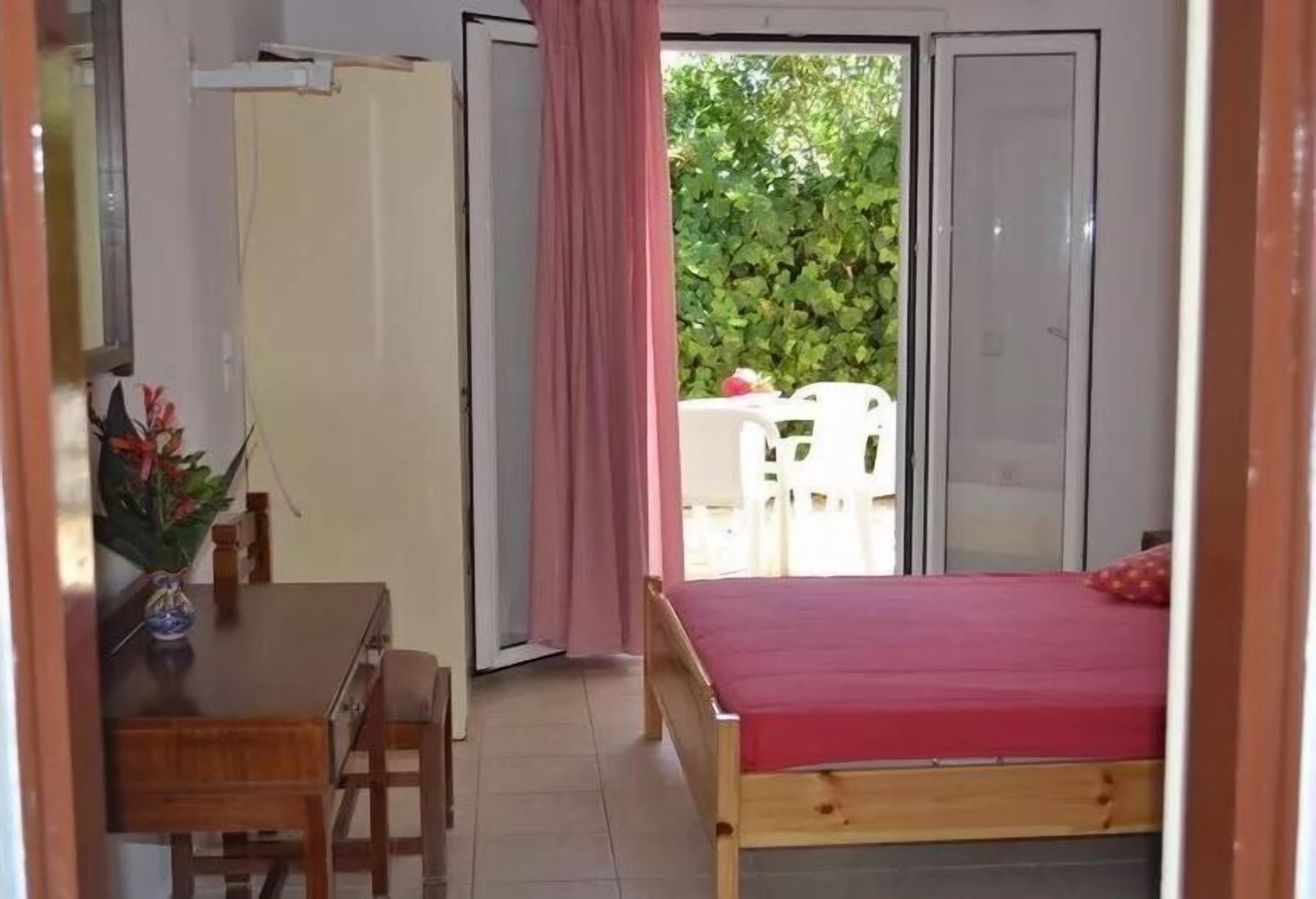 A room with a bed, a desk, and a chair. There is a sliding door leading to an outdoor area with a table and chairs.