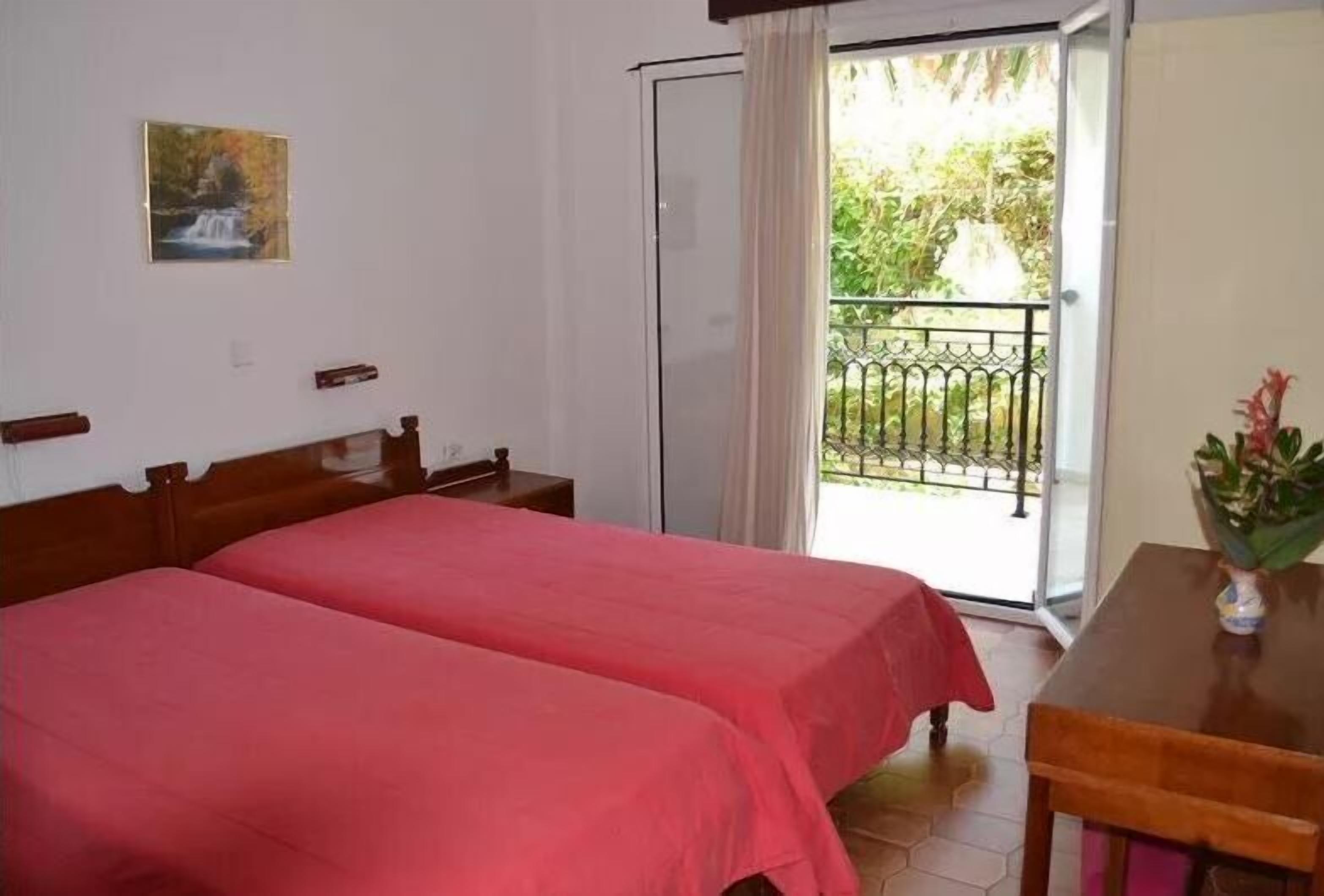 A room with two beds, a wooden table, a painting on the wall, and a balcony with a view of greenery.