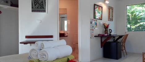 A hotel room with a bed, a desk with a chair, a small table, a mirror, and a window with a view of greenery.