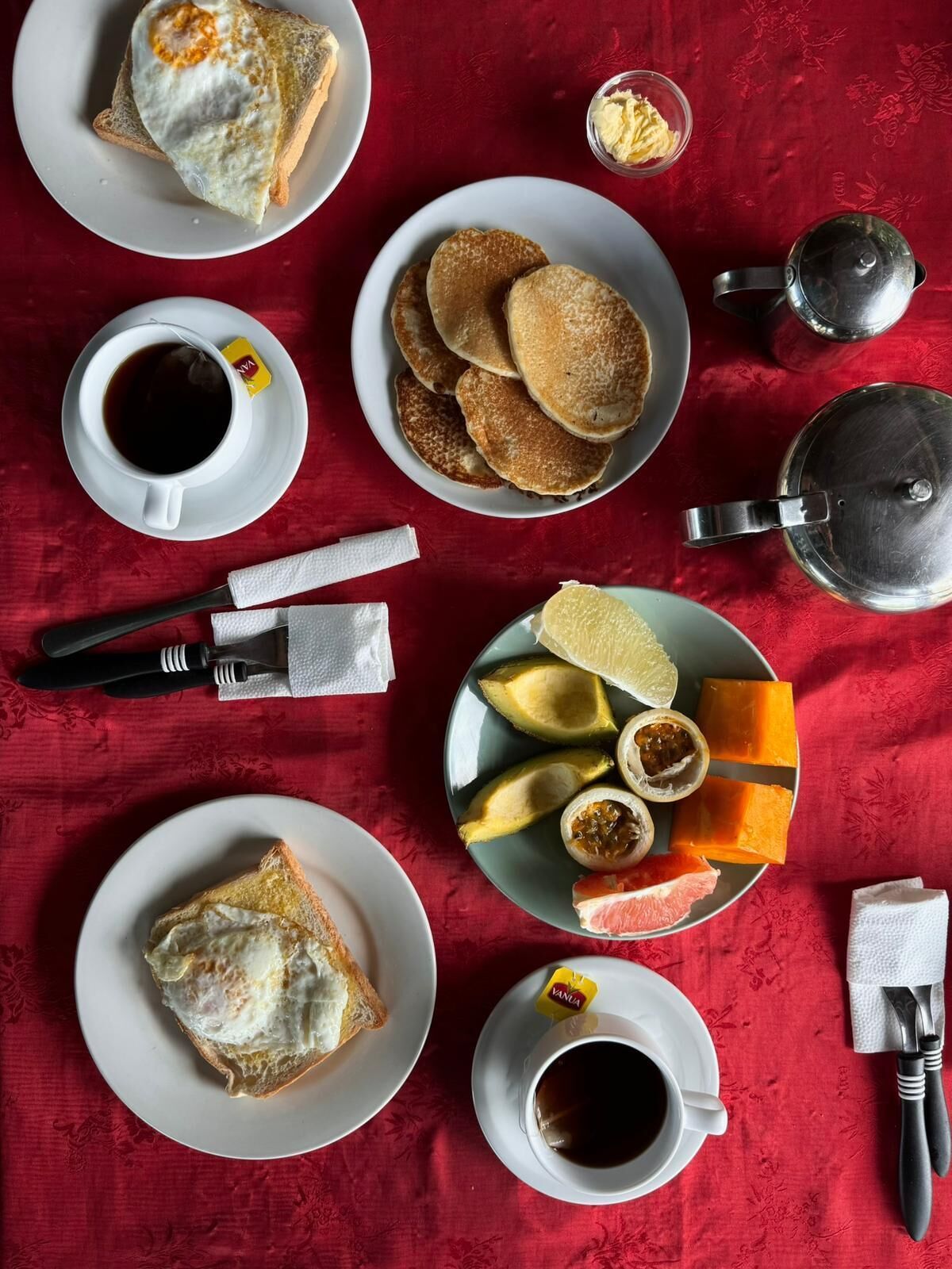 Een ontbijttafel met toast, pannenkoeken, fruit en koffie, gedekt op een rode tafelkleed.