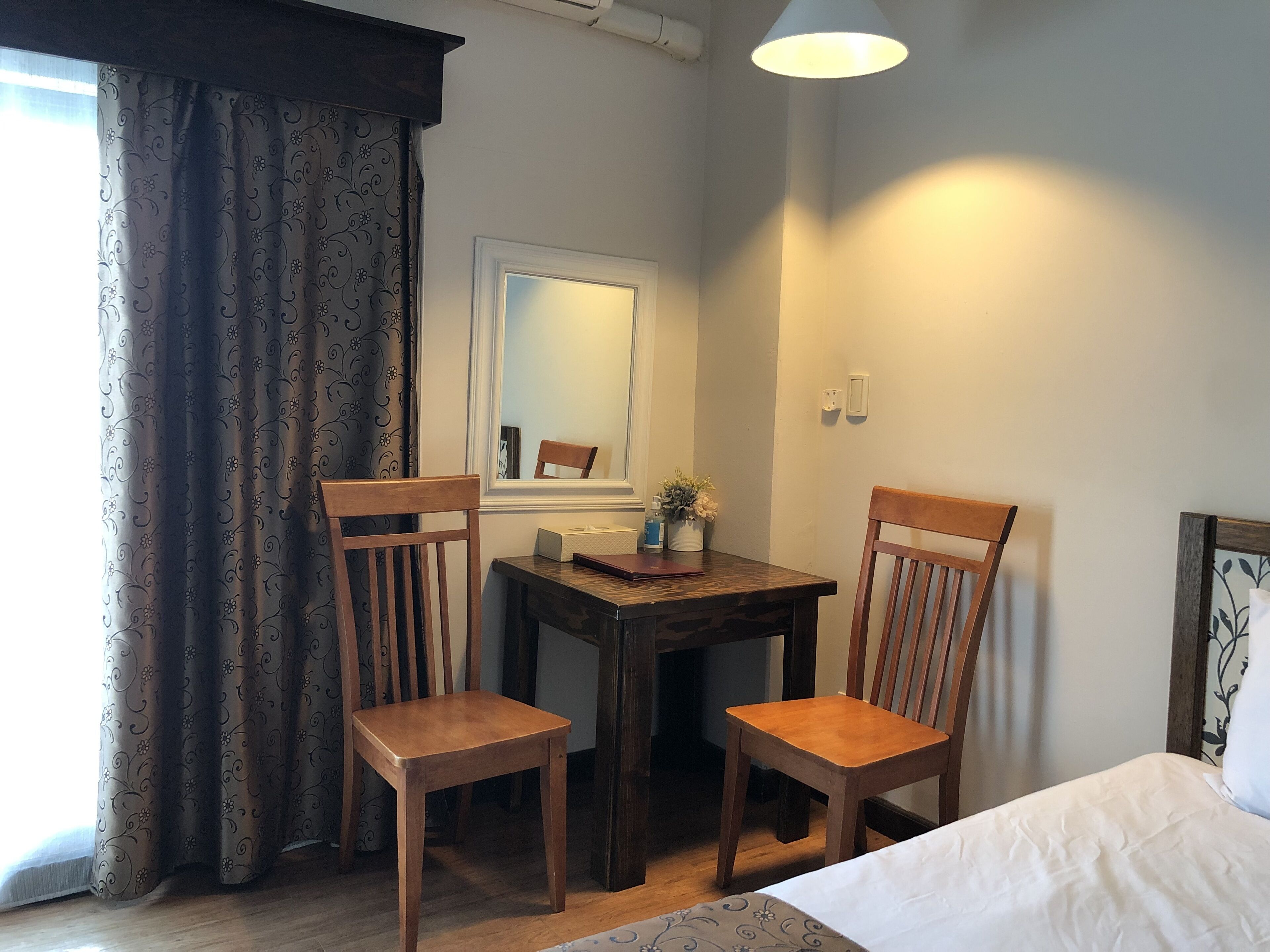 Quarto com duas cadeiras de madeira, uma mesinha, uma cama, uma janela com cortinas, um espelho e um abajur.