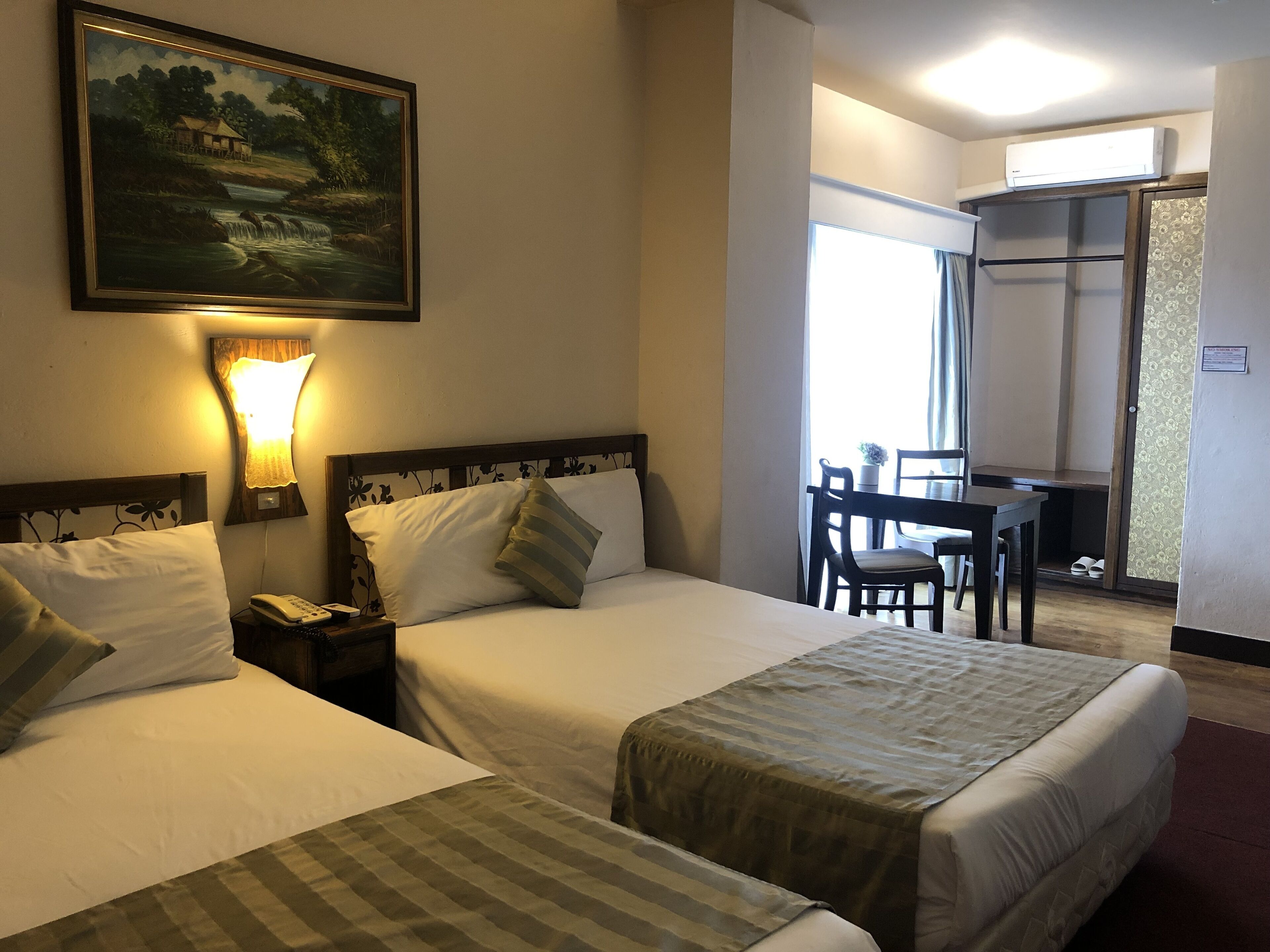 A hotel room with two beds, a desk, and a chair. There is a painting on the wall and a window with curtains.