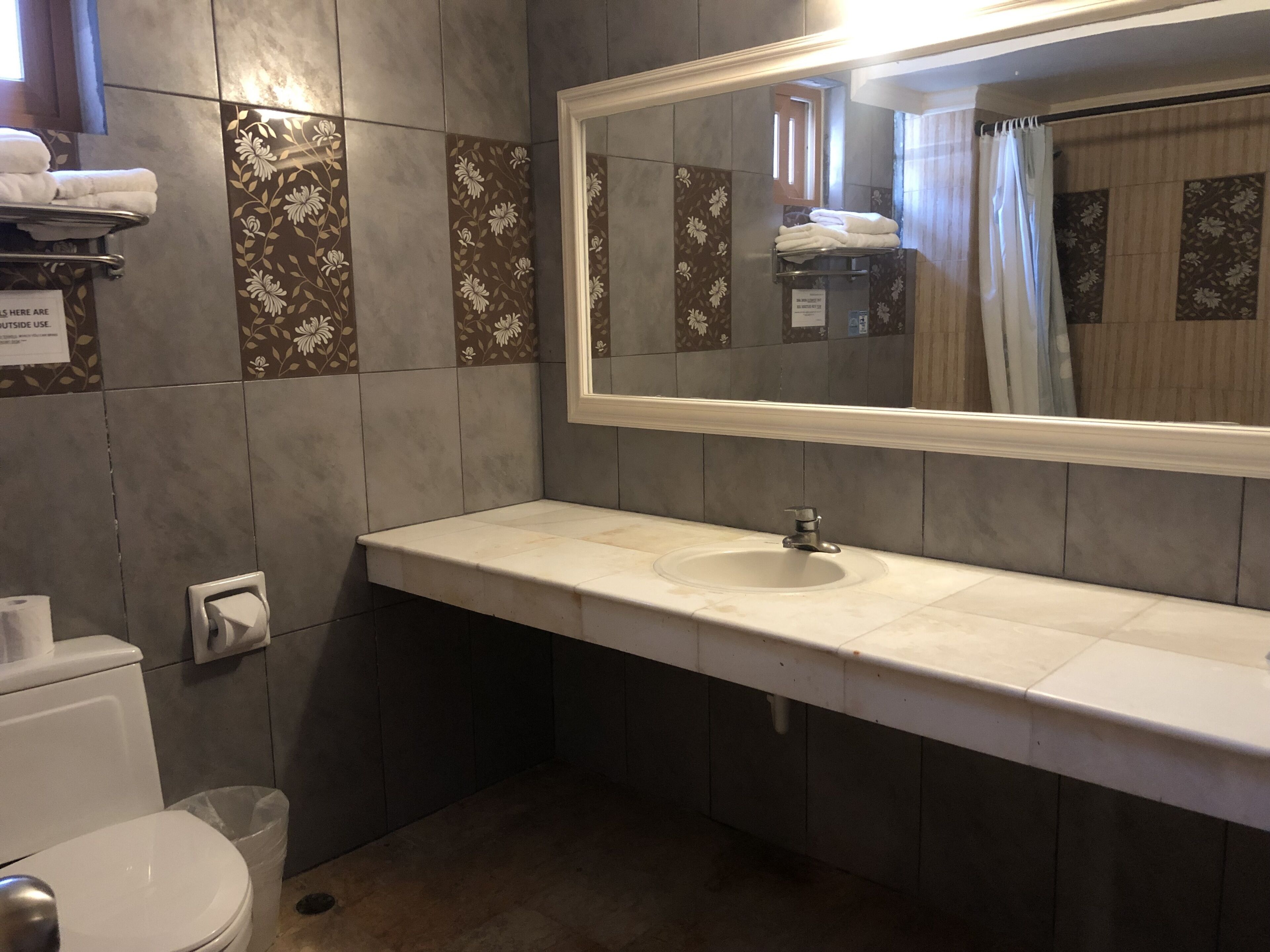Ein Badezimmer mit Waschbecken, Spiegel und Toilette. Handtücher hängen an einem Wäscheständer und ein Papierkorb steht bereit.