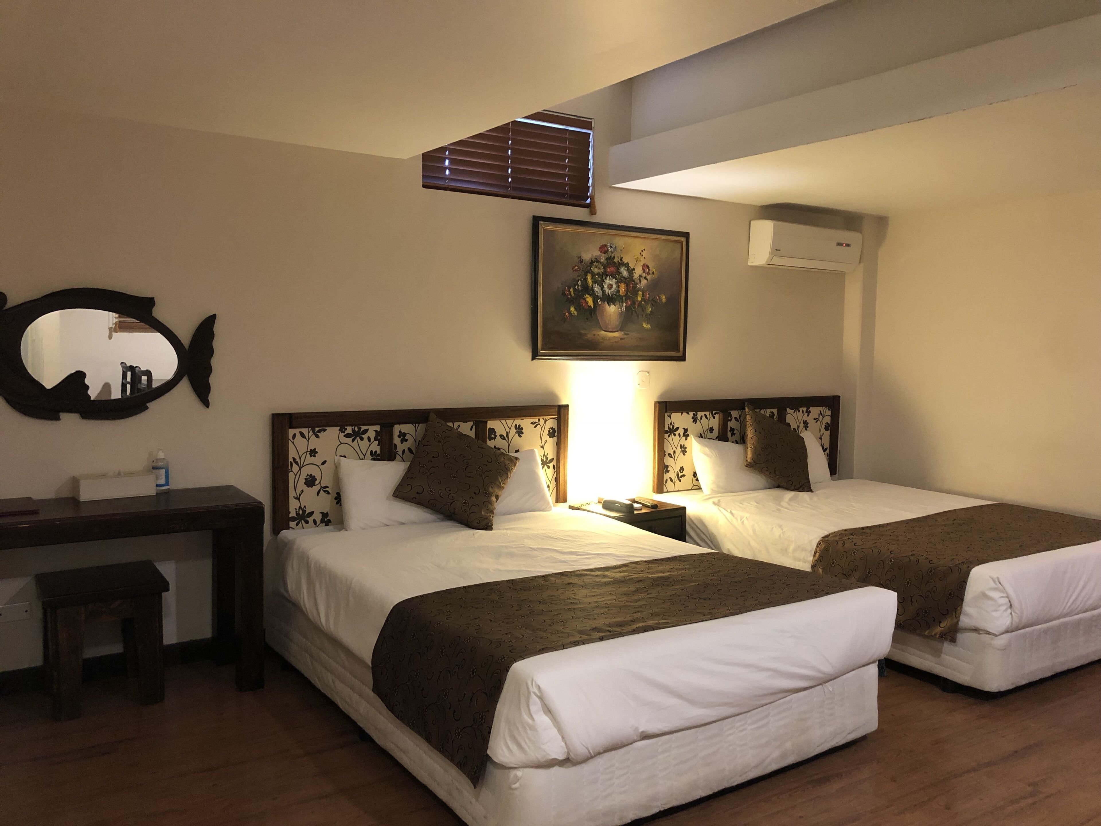 A hotel room with two beds, a desk, a mirror, a painting, and an air conditioning unit.