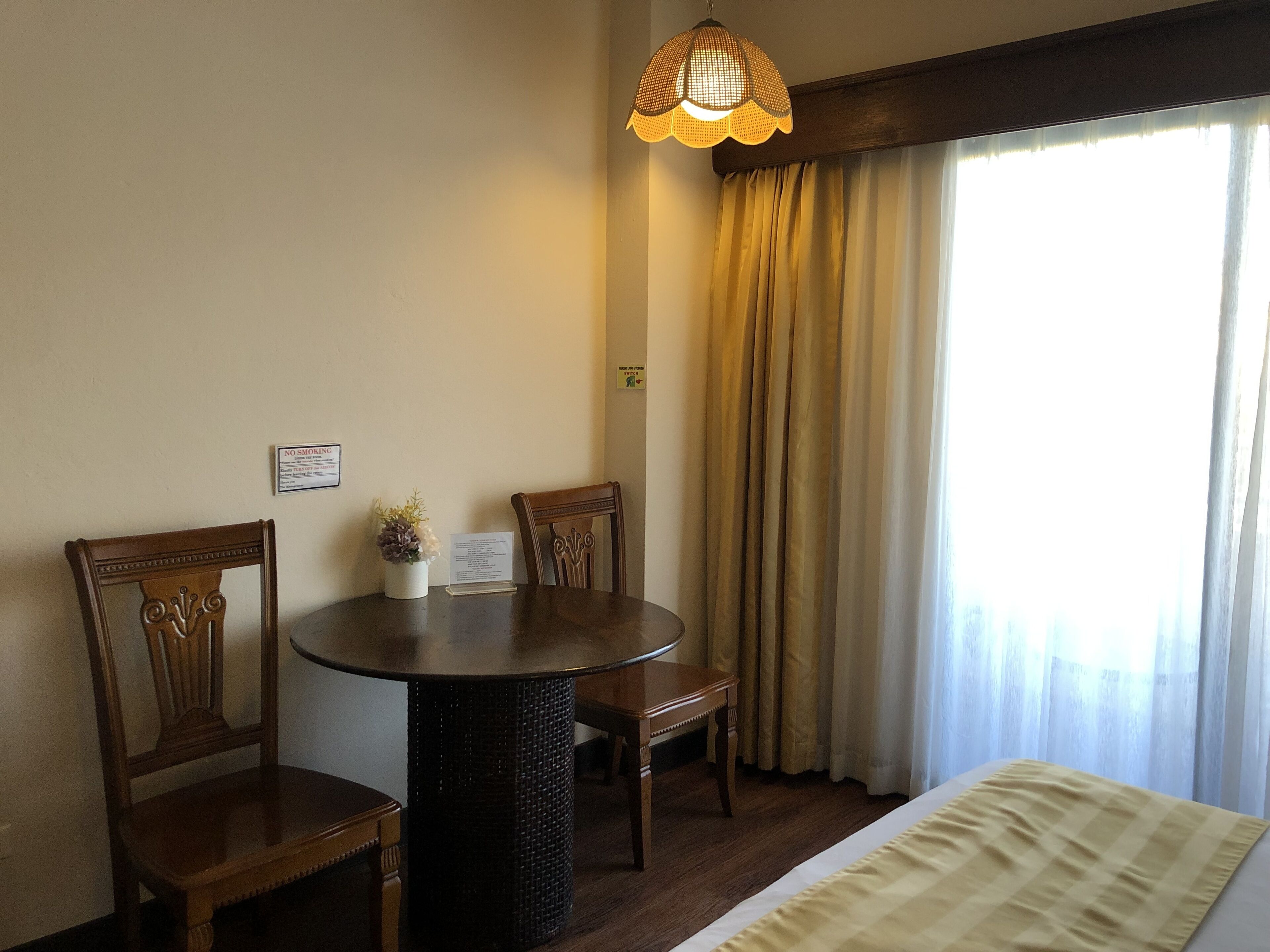 A hotel room with a round dining table, two wooden chairs, a small potted plant, a bed with a white comforter, and a window with curtains.