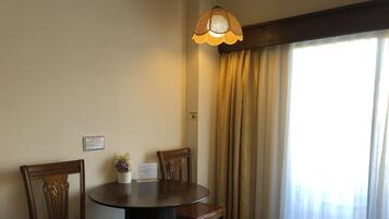 A hotel room with a round dining table, two wooden chairs, a small potted plant, a bed with a white comforter, and a window with curtains.