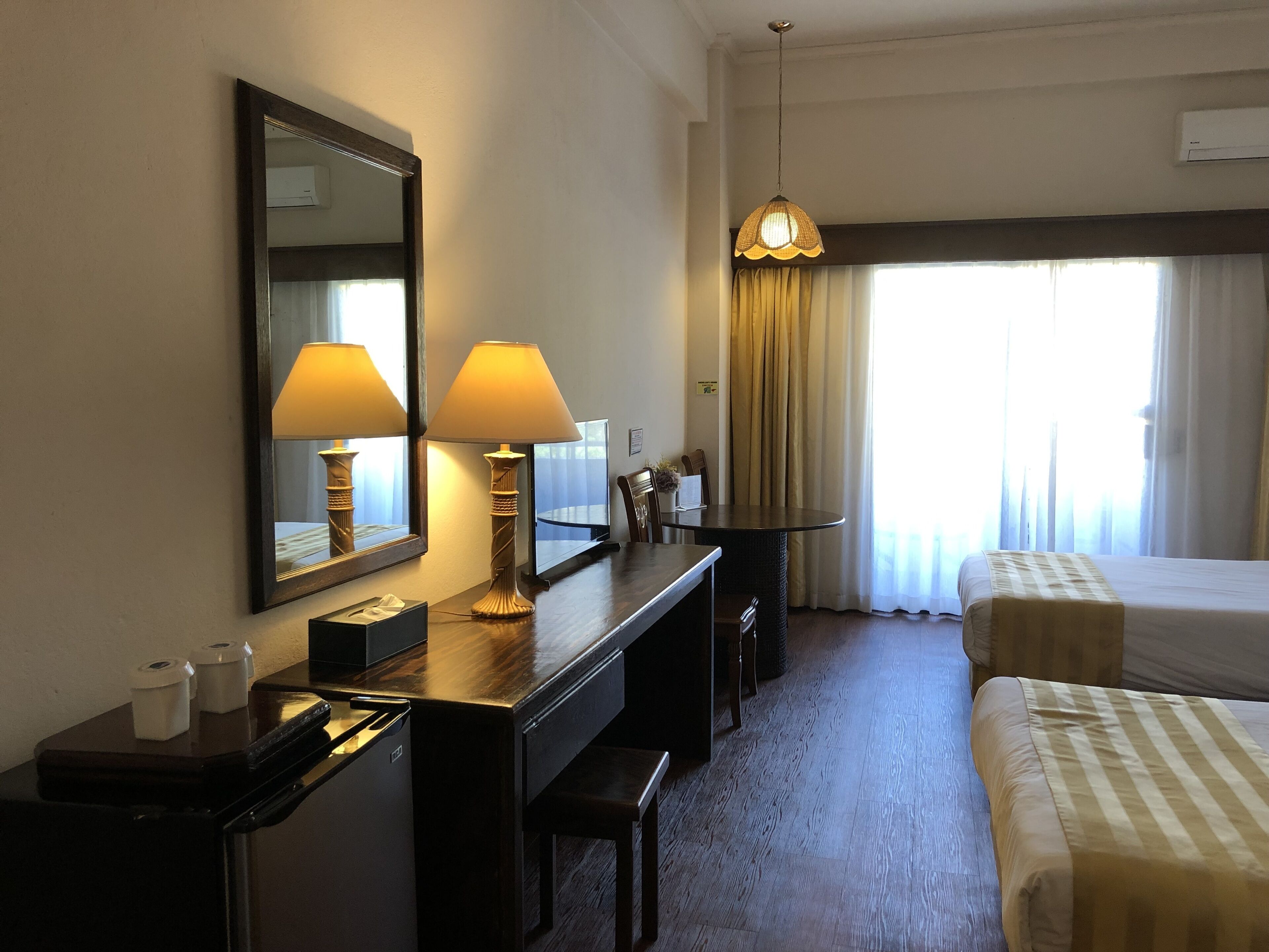 Habitación de hotel con cama, escritorio con dos lámparas, espejo, mesa de comedor y ventana con cortinas.