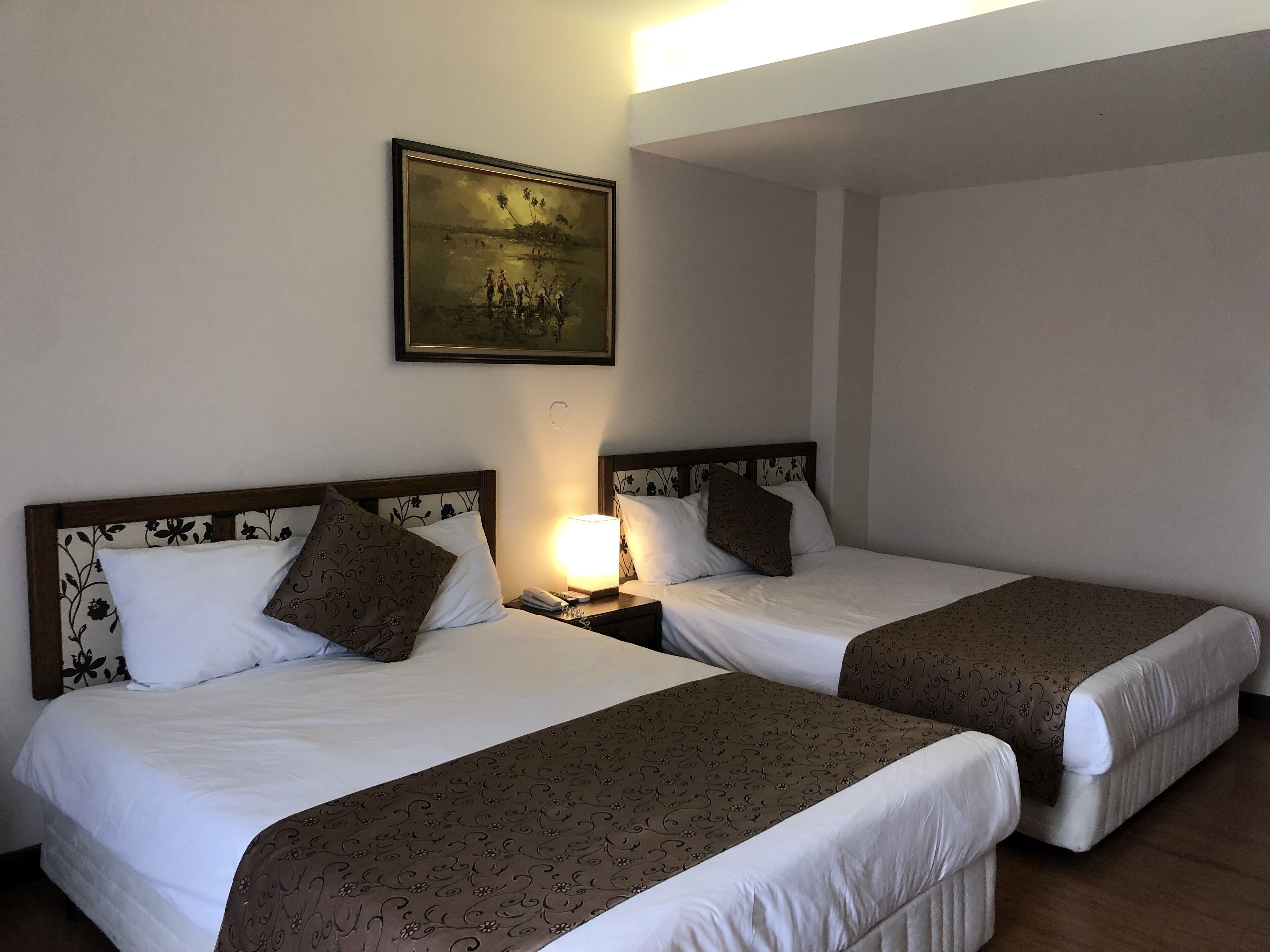 Habitación de hotel con dos camas, cada una con cabecero, mesita de noche con lámpara y un cuadro enmarcado en la pared.