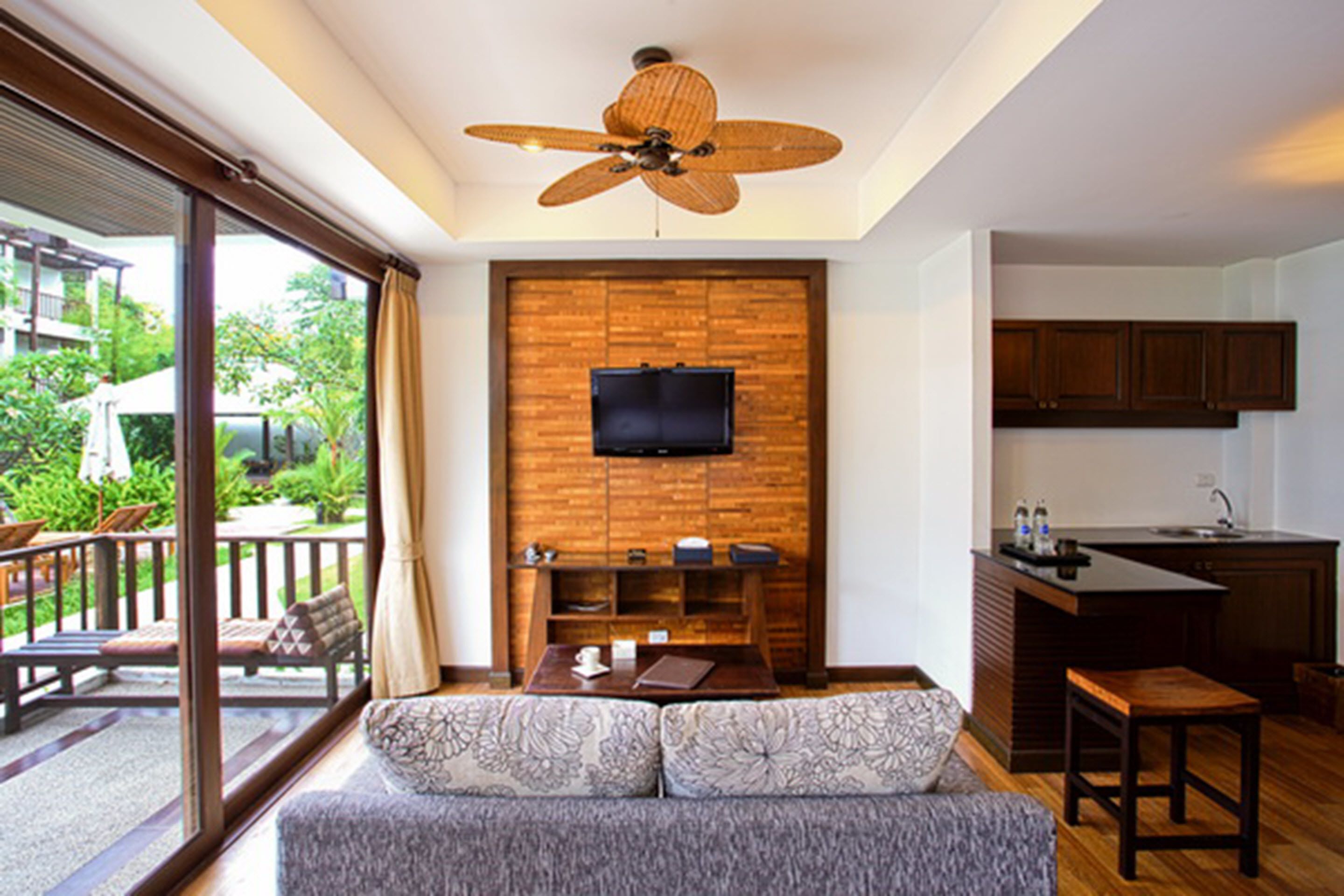 Una sala moderna con un sofá, una mesa de centro y un televisor montado en una pared de madera. Se ve un balcón con muebles de exterior a través de amplios ventanales.