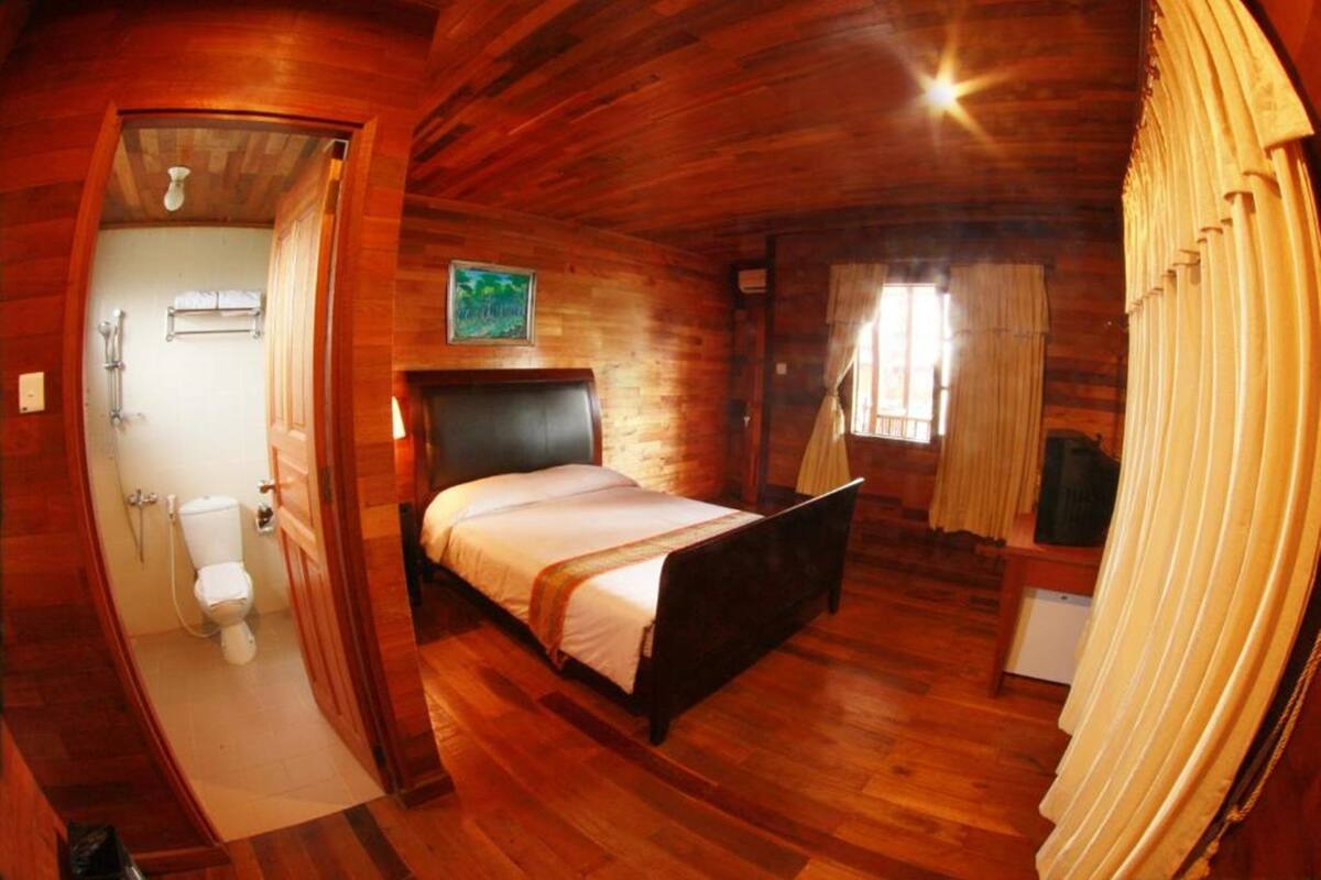 A wooden cabin bedroom with a bed, a small bathroom, and a window with curtains.