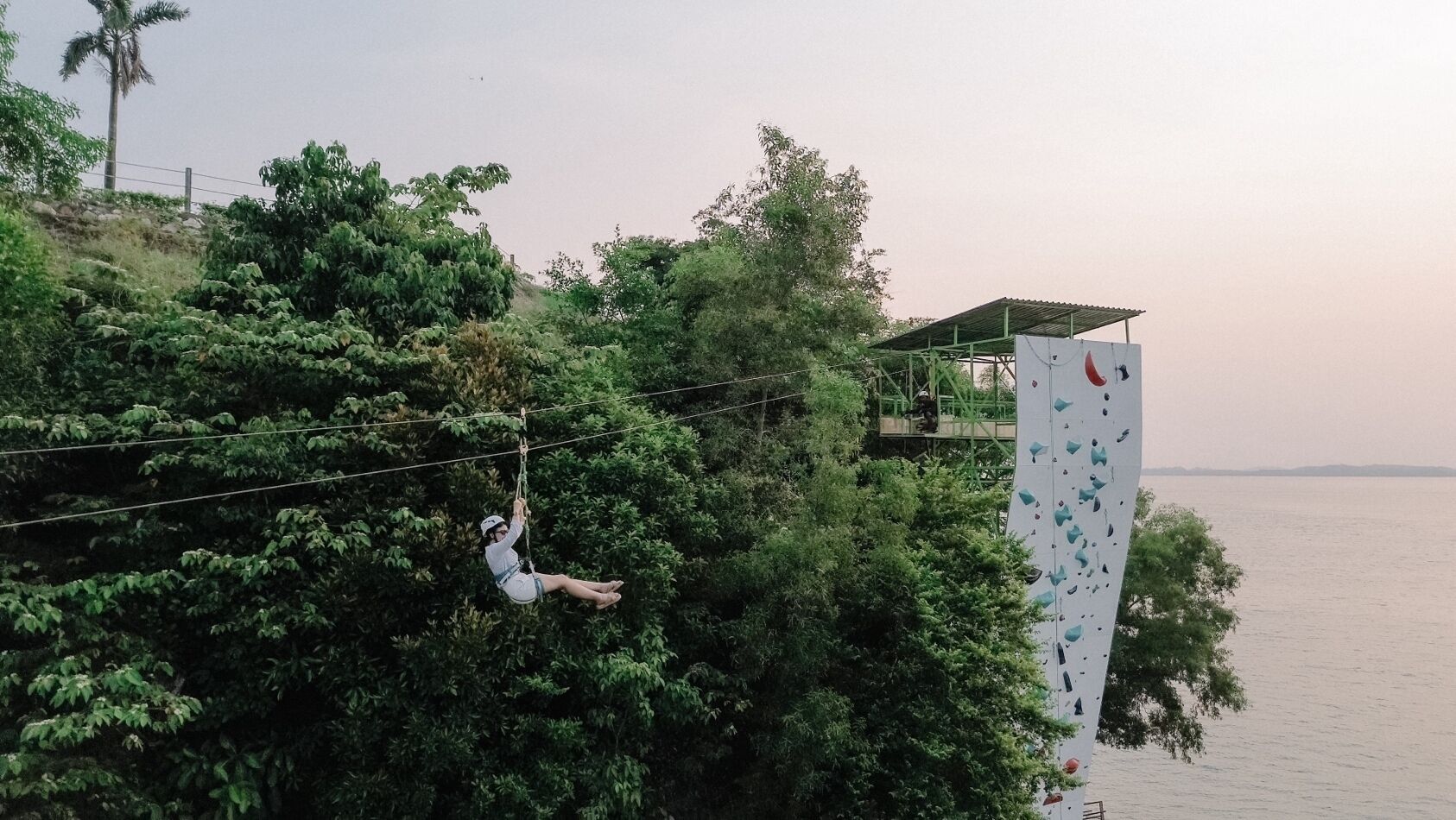 A person zip-lining over a lush green area with a building and water in the background.