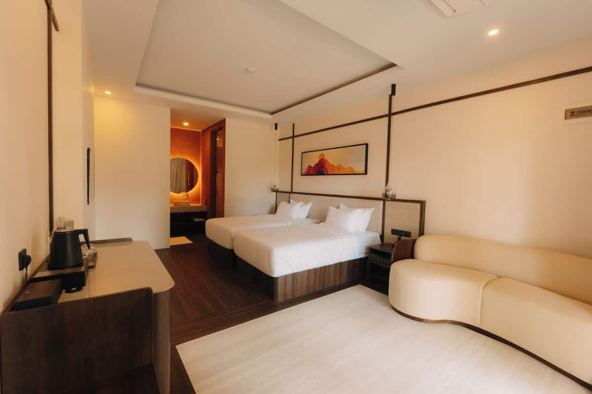 A hotel room with two beds, a desk, a sofa, a painting, and a view of the interior.