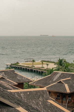 A coastal view with a pier leading to a beach area, wooden buildings with thatched roofs, and a ship in the distance.