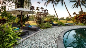 Un espace aménagé au bord de la piscine, avec des chaises longues et des parasols, entouré de plantes tropicales et de palmiers.
