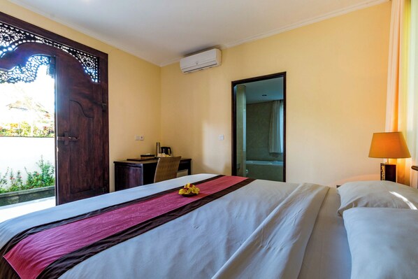 A hotel room with a bed, a desk, a chair, an air conditioning unit, and a window with a view of greenery. - Villa Taman di Blayu (Tabanan)
